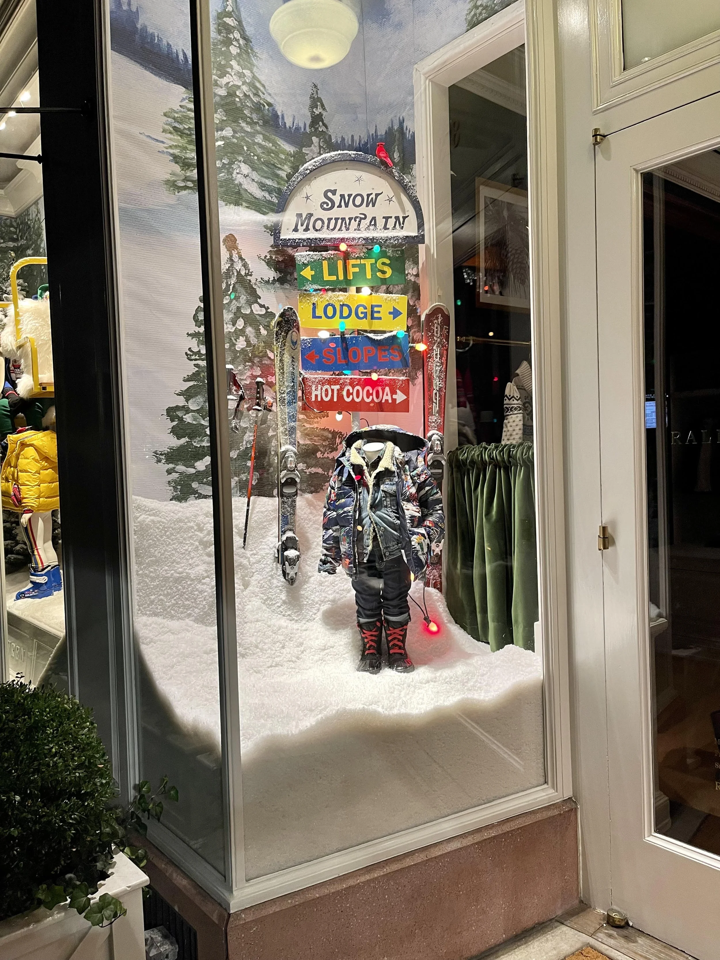 Winter themed storefront display with a snow-covered ground, a small mannequin dressed in winter gear, vintage skis, and a sign with directions to lifts, lodge, slopes, and hot cocoa, along with snow-covered trees and a painted mountain background.