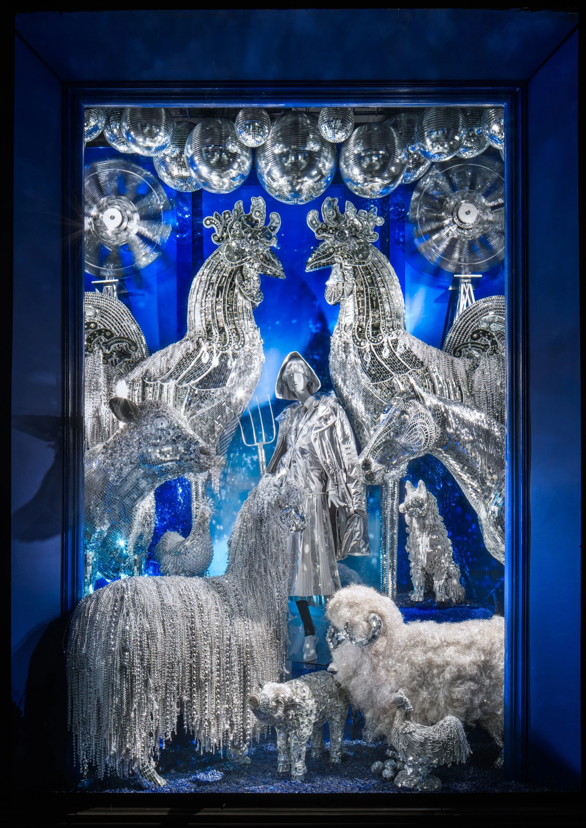 A window display featuring metallic and crystal animals, a woman in silver clothing, and large reflective disco balls, set against a blue background.