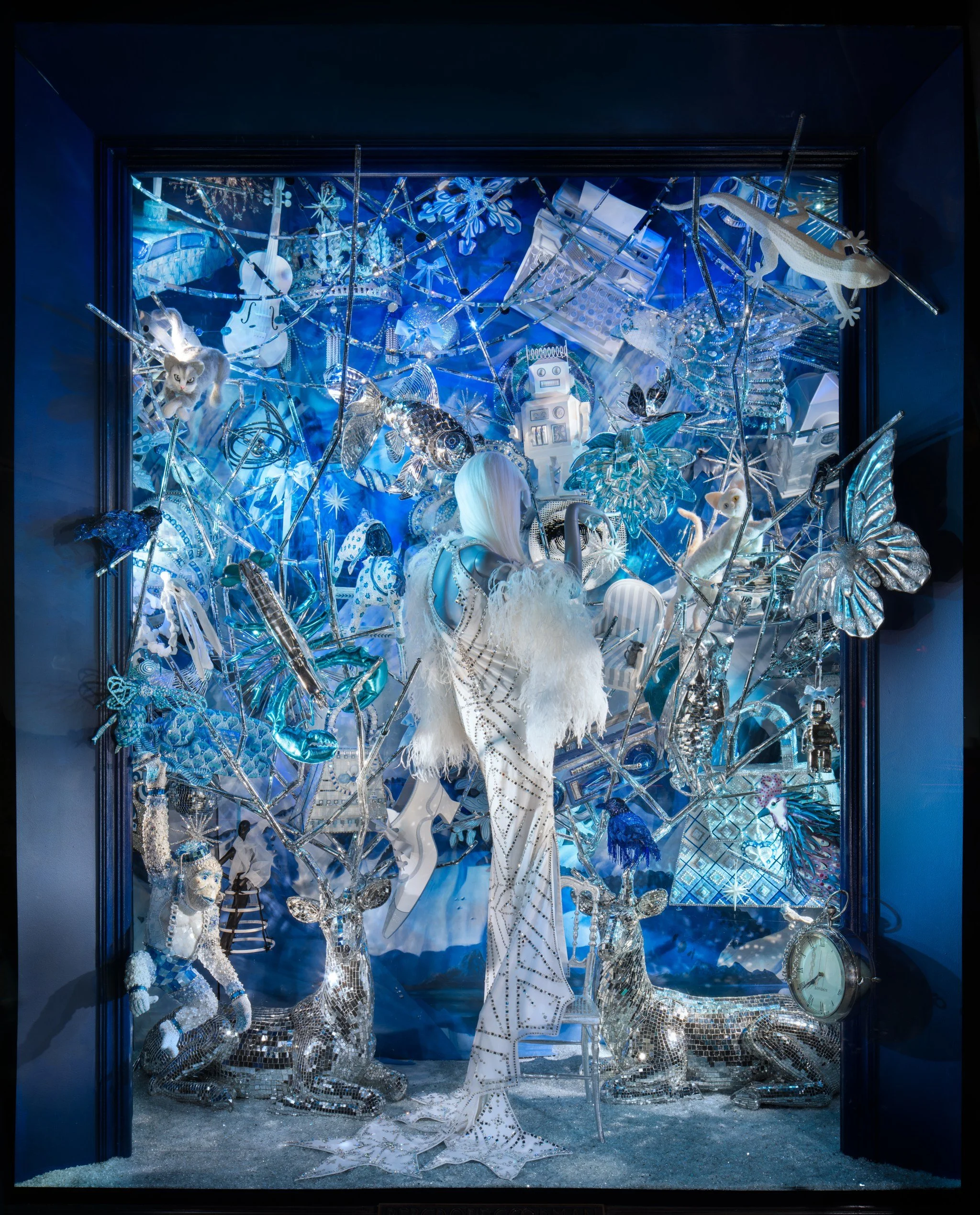 A festive window display decorated with Christmas-themed items in silver, white, and blue. Features a mannequin with long white hair in a white outfit, surrounded by shiny reindeer, silver butterflies, and small animals. The scene includes snowflakes, a clock, and various sparkly ornaments.