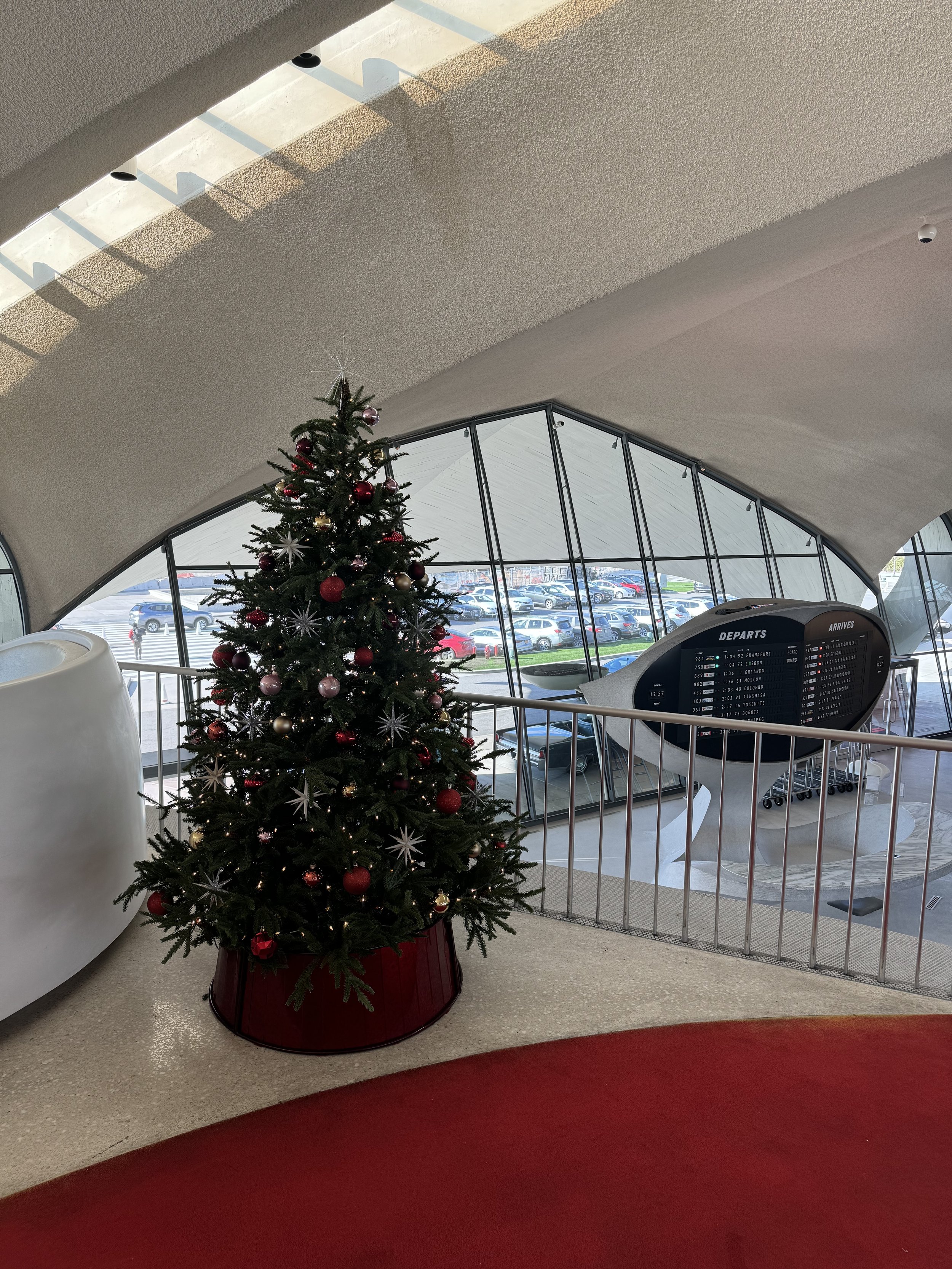 Christmas tree decorated with red and pink ornaments inside an airport terminal near a window.
