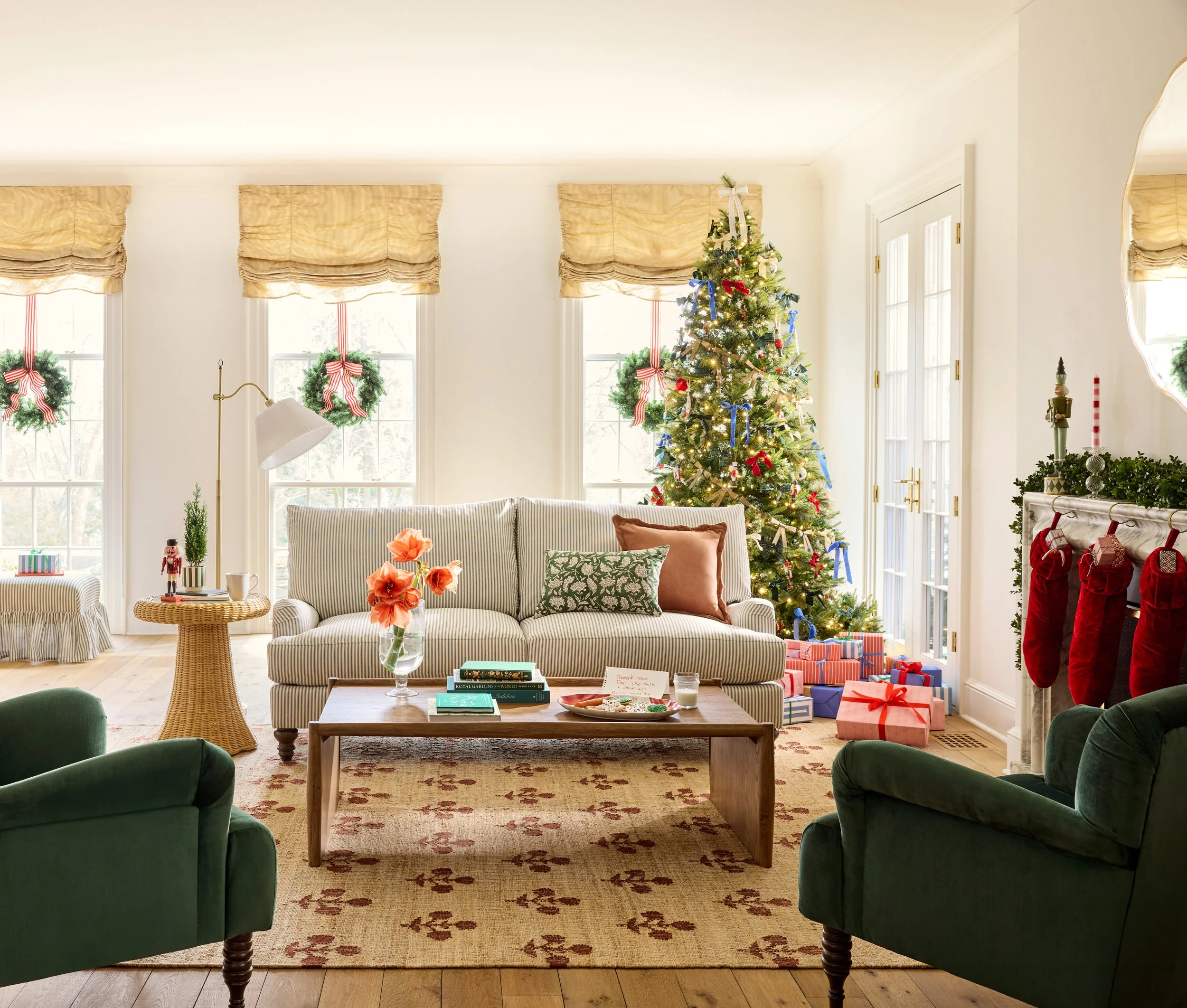 Living room decorated for Christmas with a decorated Christmas tree, wrapped presents, a white sofa with decorative pillows, green armchairs, a mirror, a fireplace with stockings, and windows with wreaths.