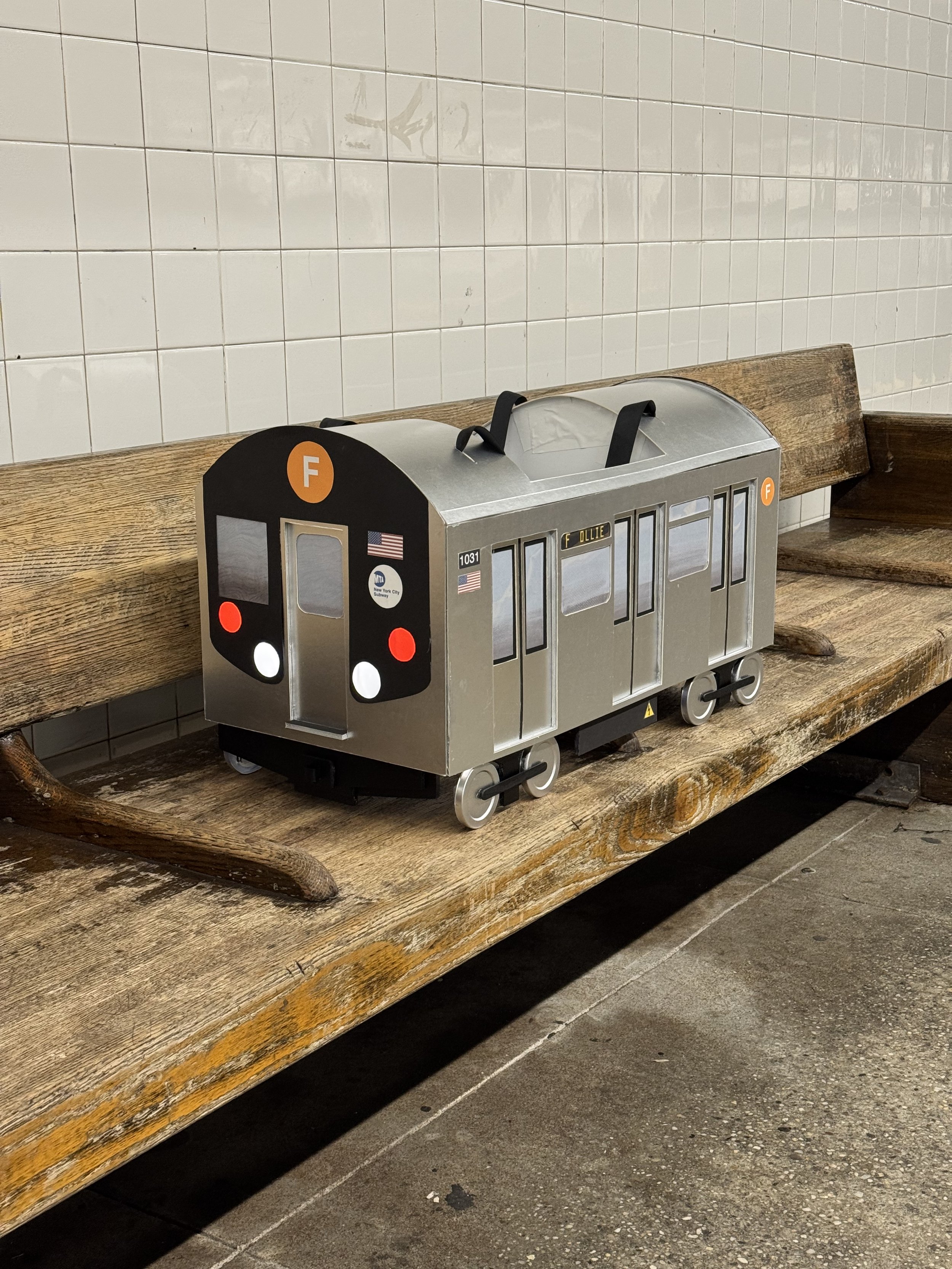 A subway train Halloween costume sitting on a wooden bench against a white tiled wall.