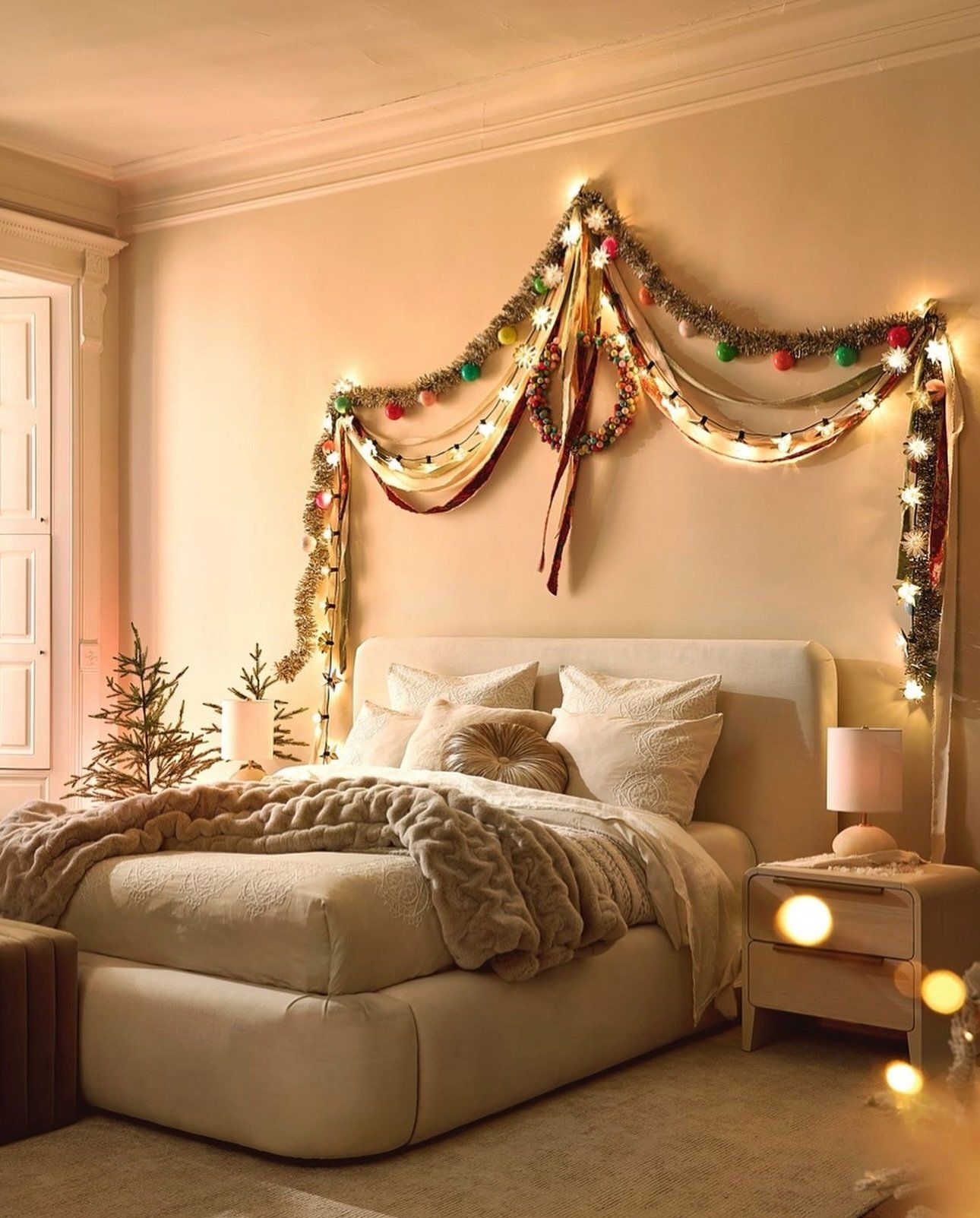 A cozy bedroom decorated for Christmas with string lights and colorful ornaments hung above the bed. The bed has white bedding, a textured blanket, and multiple pillows. Two small side tables with lamps flank the bed, and there are small Christmas trees on each side.