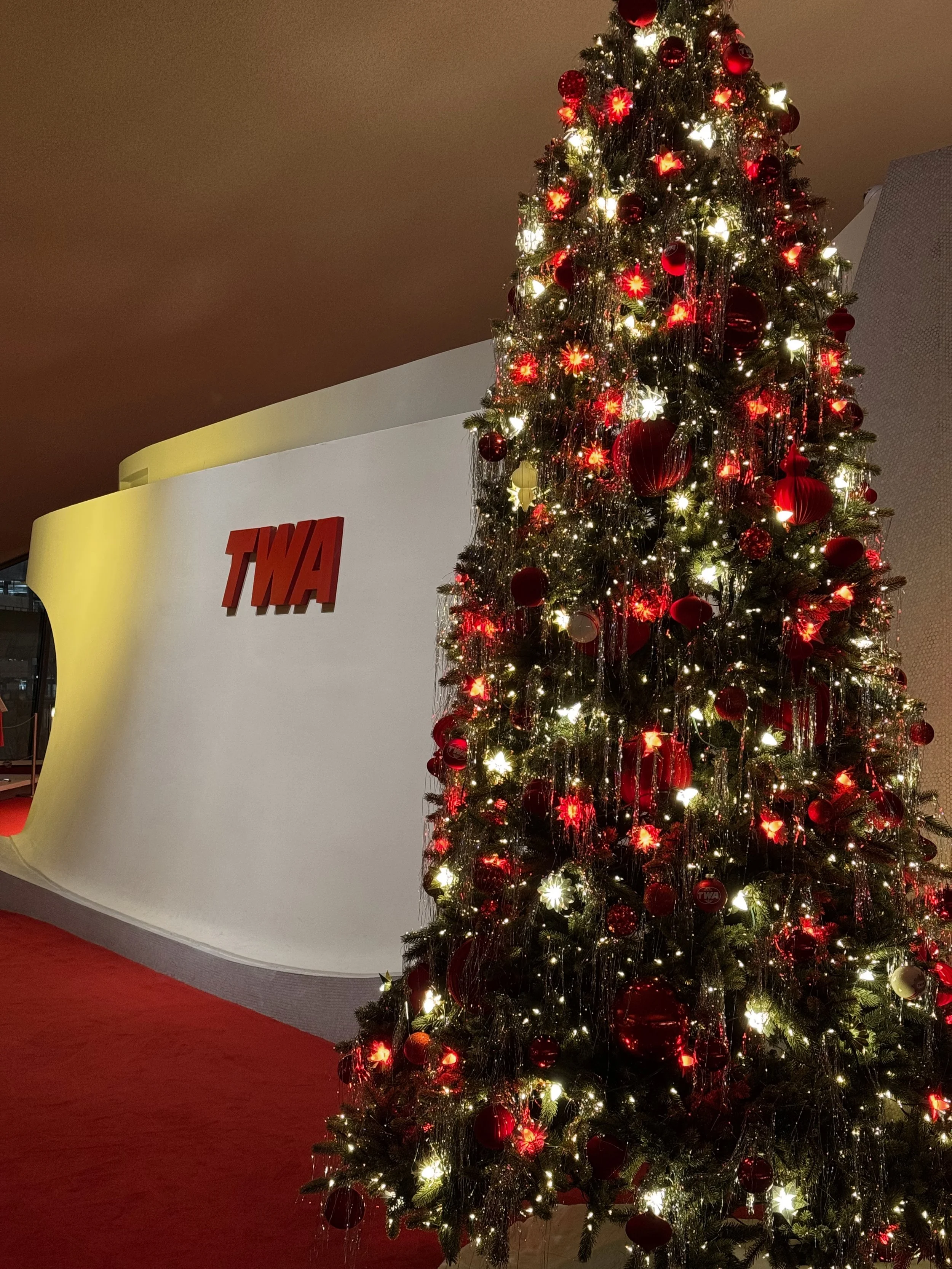 A decorated Christmas tree with red ornaments and white lights standing next to a white wall with red TWA letters in a building lobby.