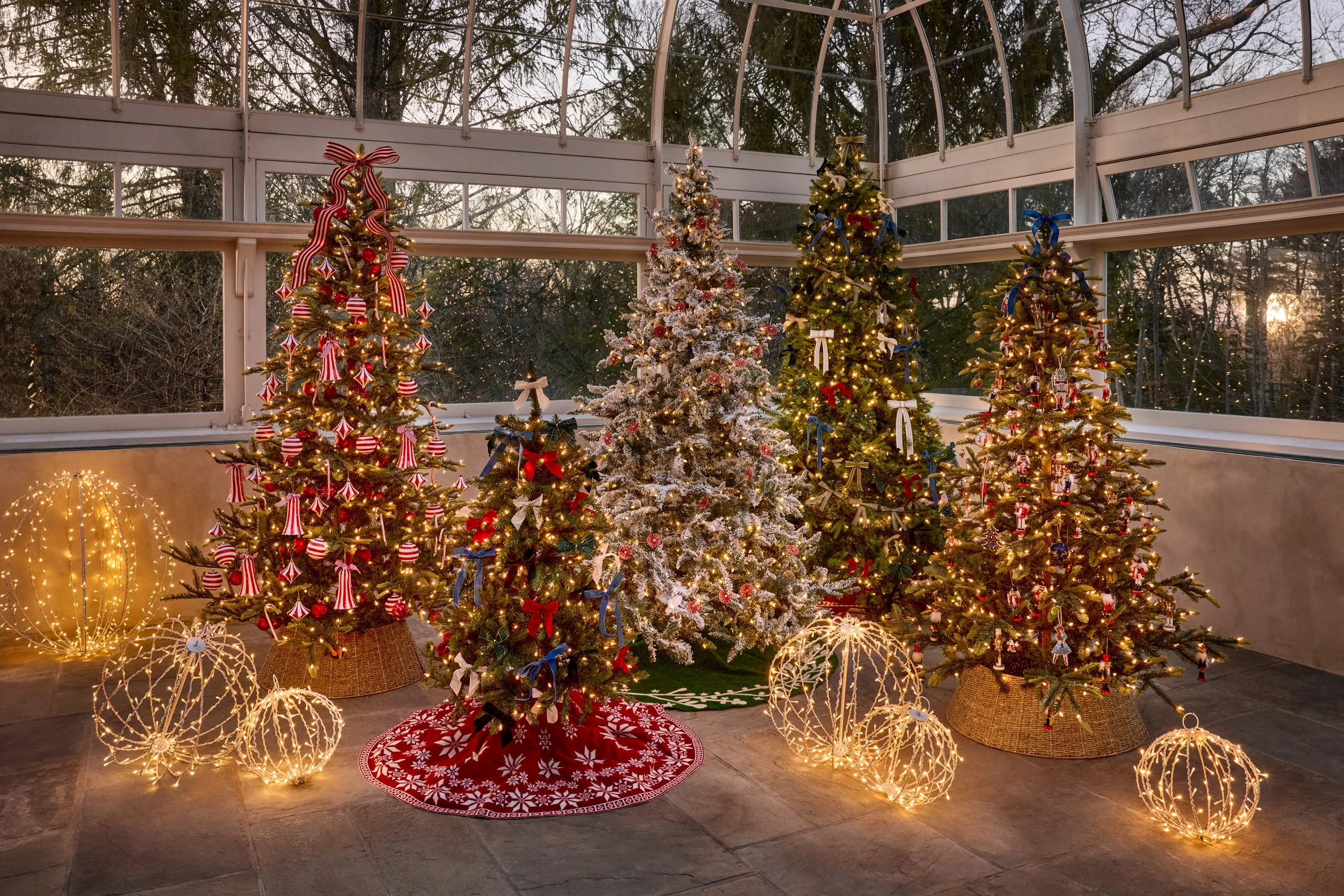 Five decorated Christmas trees inside a glass conservatory with sunlight outside, surrounded by illuminated spherical decorations on a tiled floor.