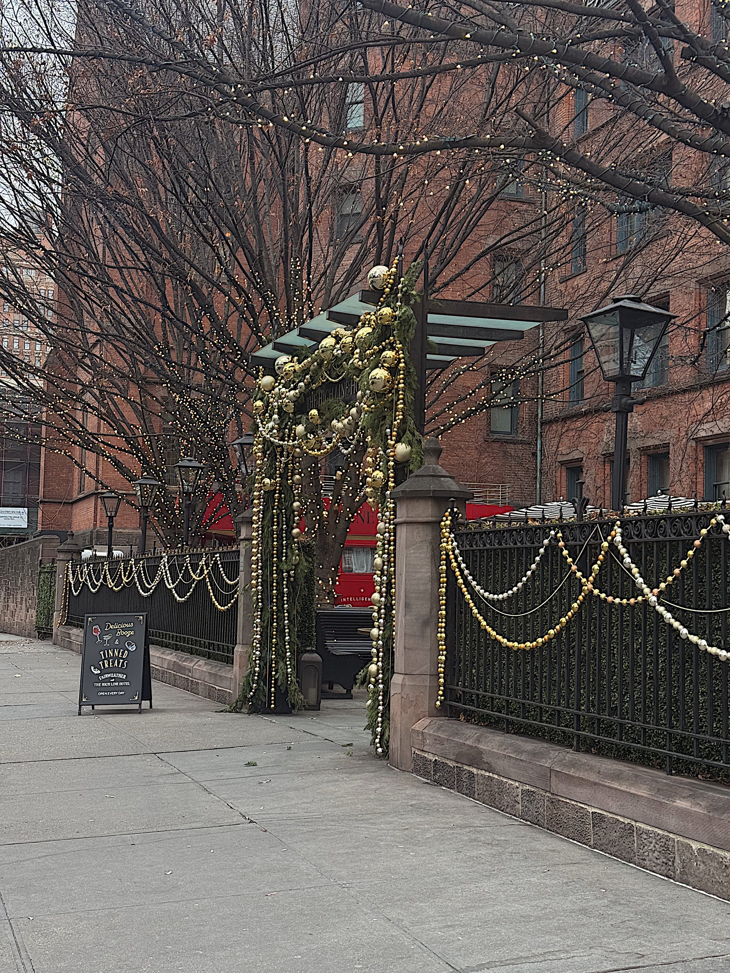 Outdoor sidewalk decorated with Christmas ornaments and lights, near leafless trees and a black metal fence, with a sign advertising baked treats.