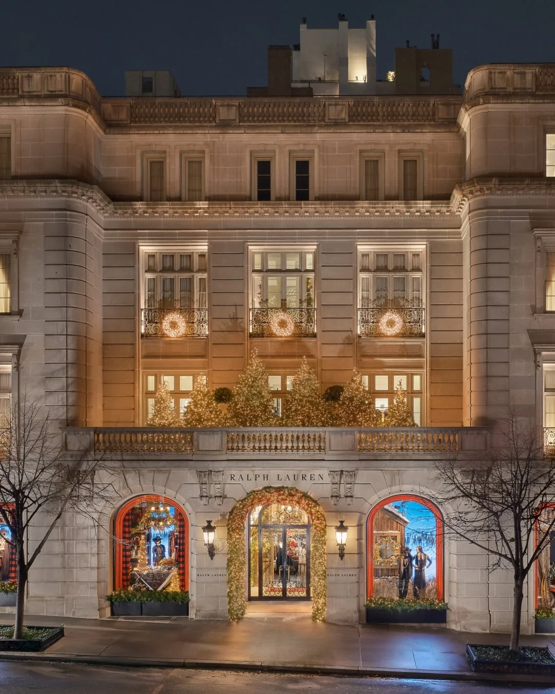 Night view of a decorated Ralph Lauren store with holiday lights, Christmas trees, and mannequins in window displays.