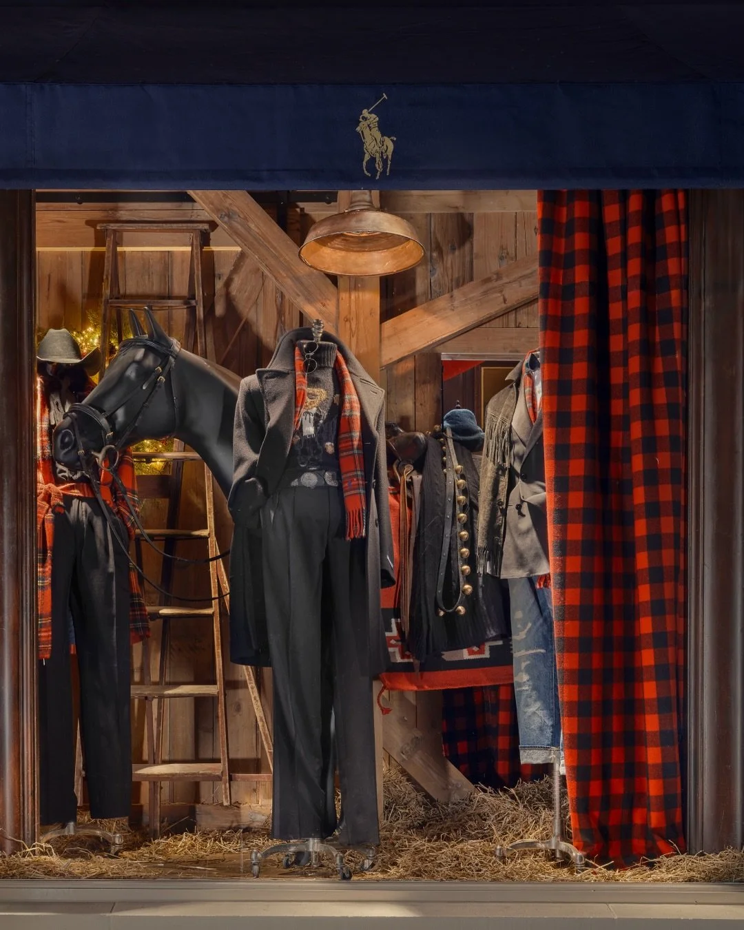 Store display with mannequins dressed in western-style clothing, including coats, scarves, and pants. The background features wooden walls, a ladder, and a curtain with red and black checkered pattern.