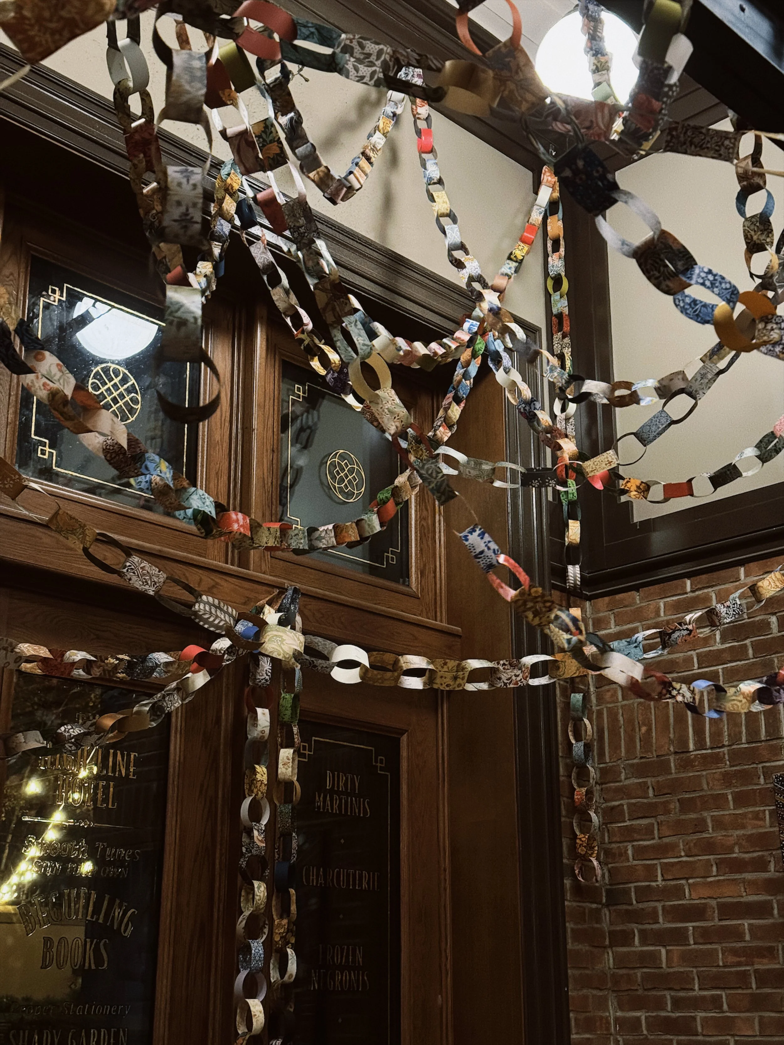 Colorful handcrafted paper chain garlands hanging from a ceiling, with a wooden-paneled wall and brick wall behind.