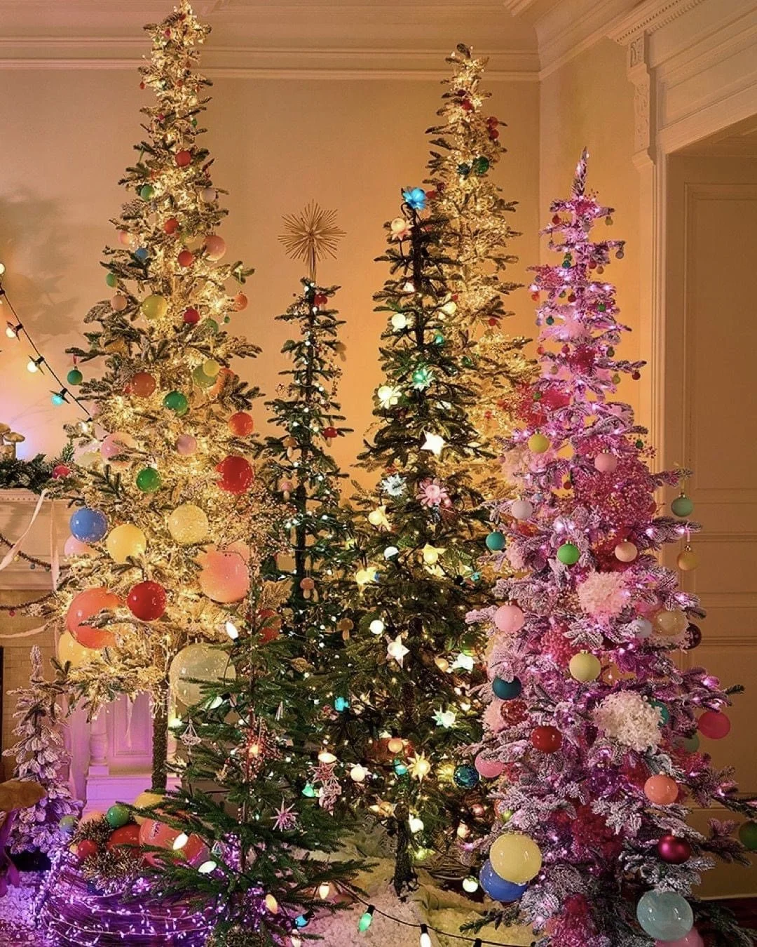Four decorated Christmas trees with ornaments and lights in a warmly lit room.