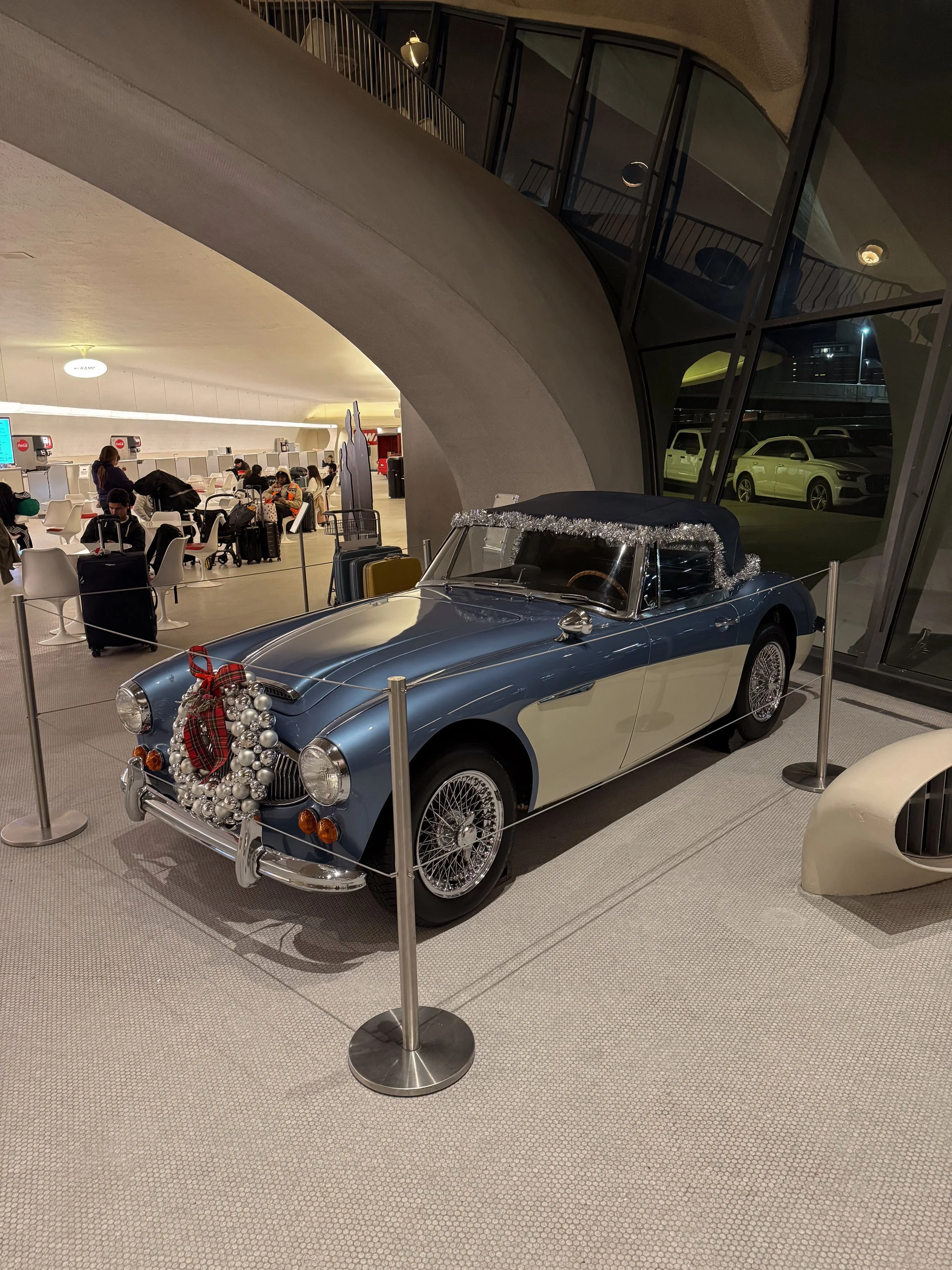 Blue vintage convertible car decorated with Christmas ornaments and tinsel, displayed at an airport terminal.