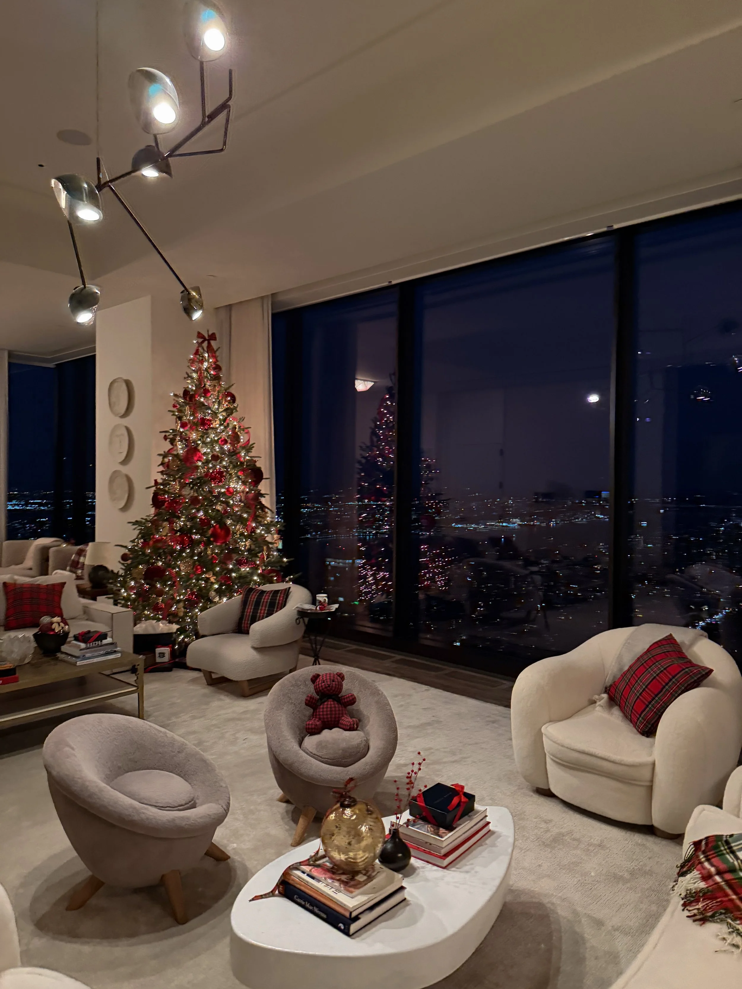 Living room with Christmas tree decorated with red and gold ornaments, presents, and a teddy bear on a chair, with large windows showing a city view at night.
