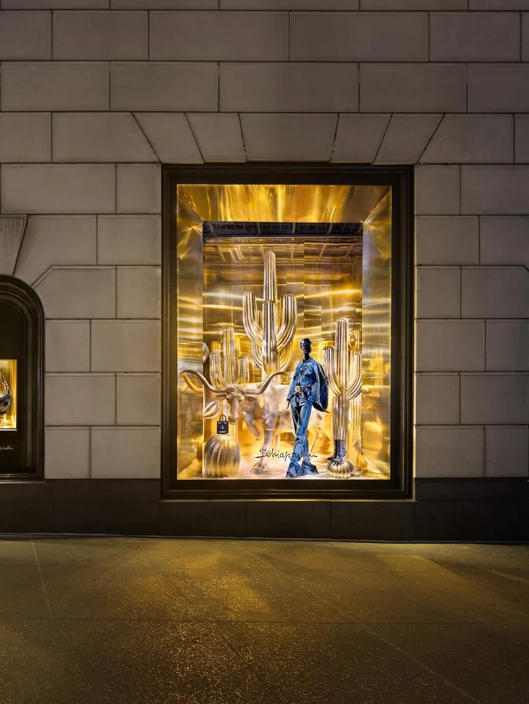 A store window display with a gold-themed desert scene featuring large cacti, a steer skull, and a model dressed in denim, set against a gold reflective background.