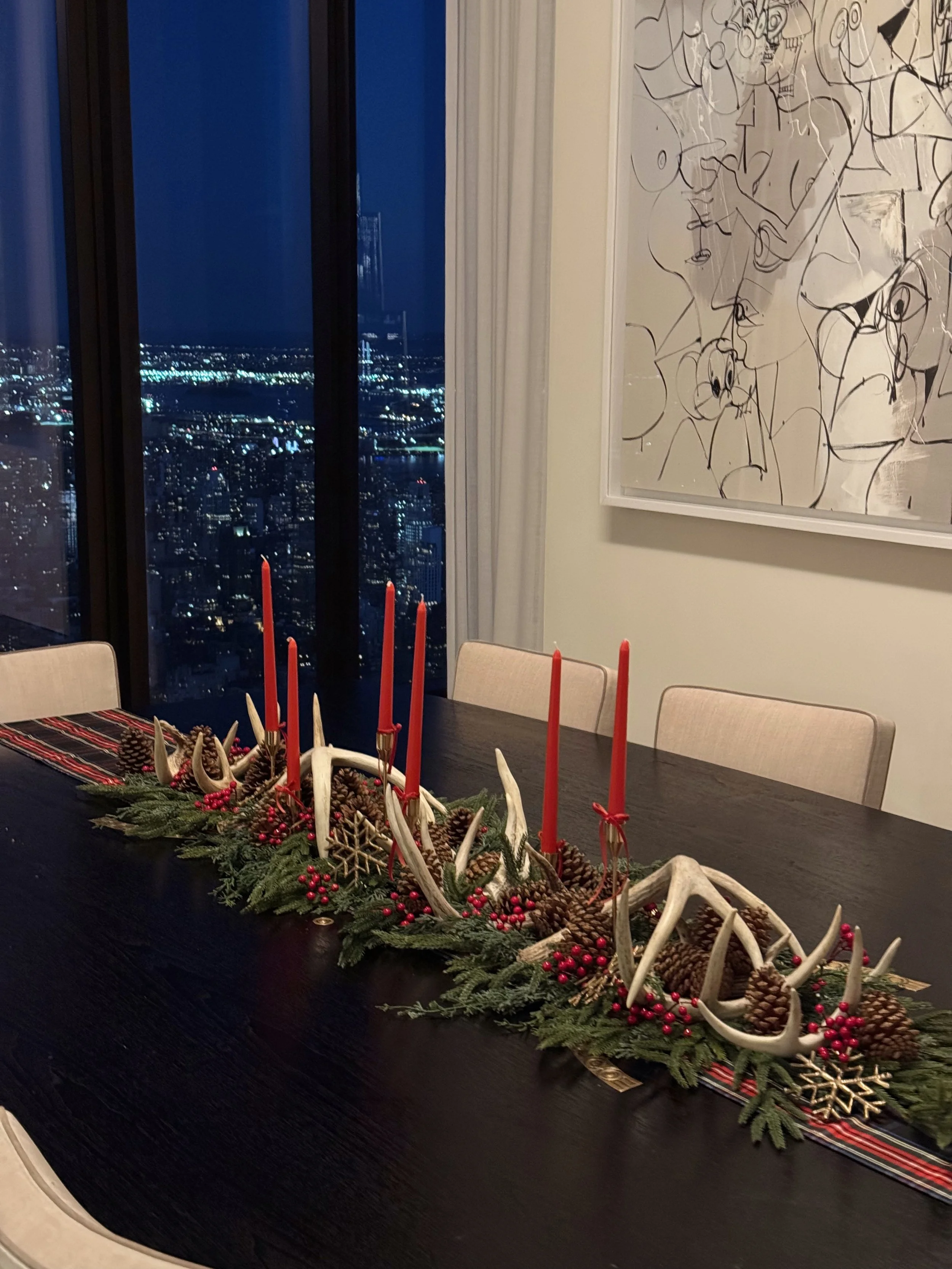 A dining table with a Christmas centerpiece featuring pinecones, red berries, and white antlers, with red candles, in a high-rise apartment with a city view at night and modern artwork on the wall.