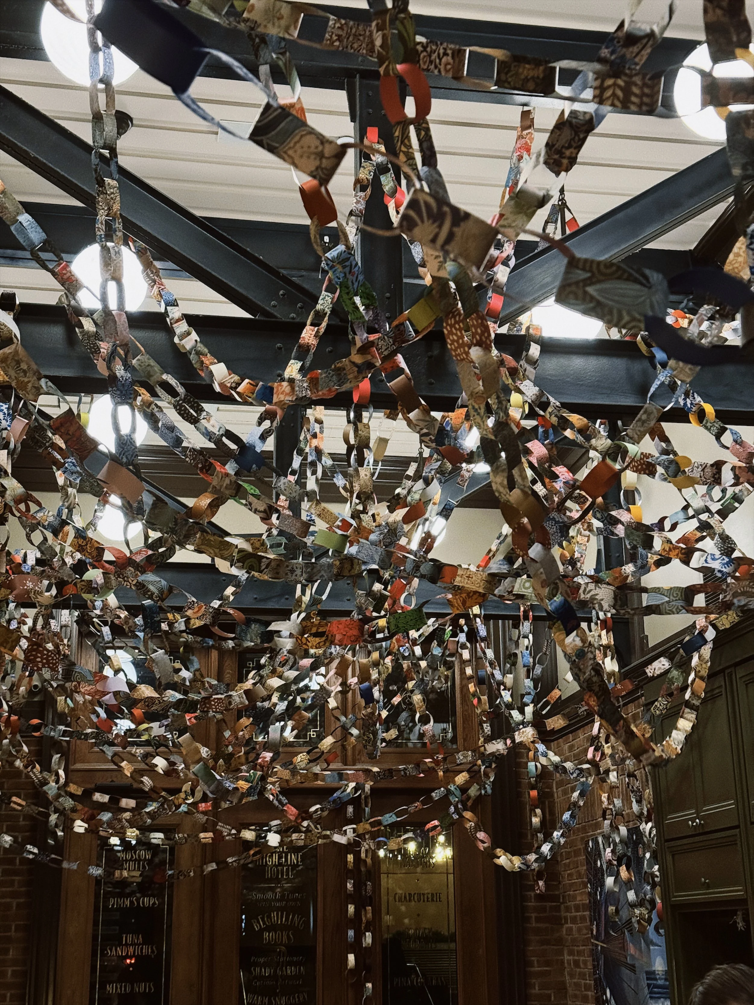 Colorful paper chains hang from the ceiling, forming an elaborate decoration in a restaurant or cafe with wood-paneled walls.