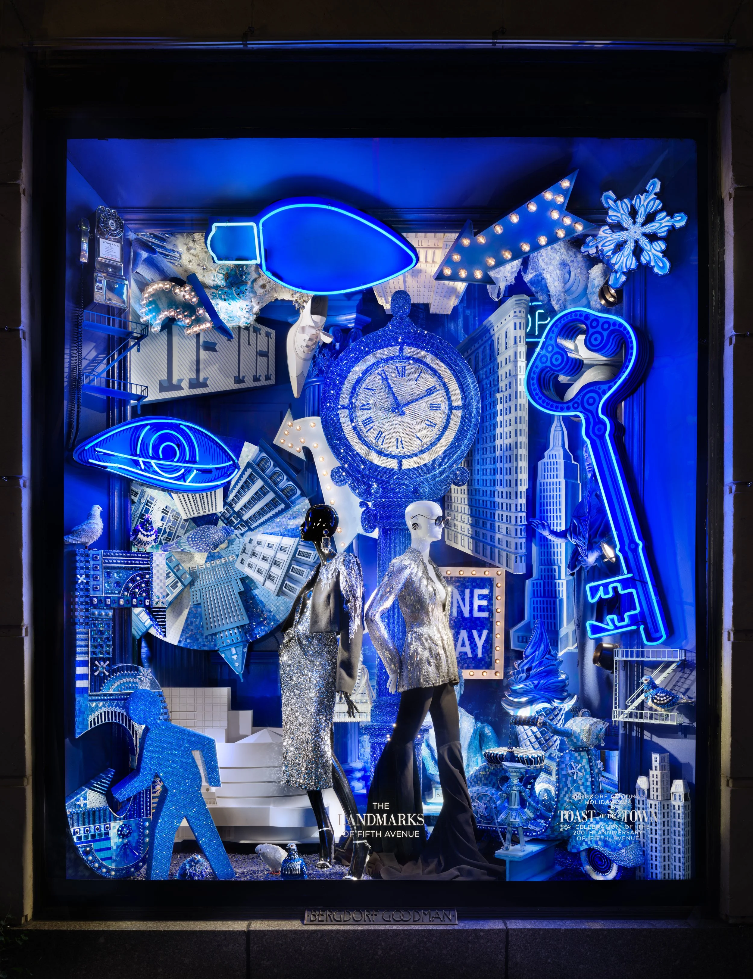 Blue-themed window display with mannequins in shiny outfits, large clock, illuminated arrows and keys, buildings, snowflake, eye, bird, and various decorative objects.