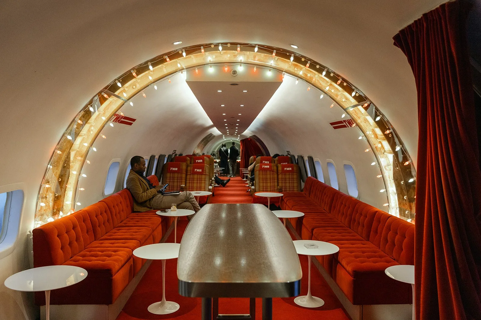 Interior of a luxurious airplane cabin with red velvet seating along the walls, small white tables, and a large table in the center. Passengers are seated and walking in the background, with a decorative light arch overhead and red curtains on the side.
