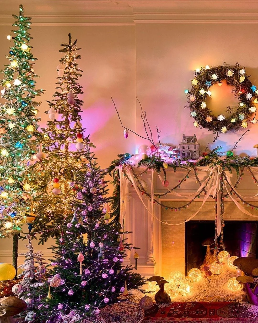 Festively decorated living room with Christmas trees, a wreath, and string lights. The fireplace mantel and floor are adorned with greenery, ornaments, and glowing orbs, creating a warm holiday atmosphere.