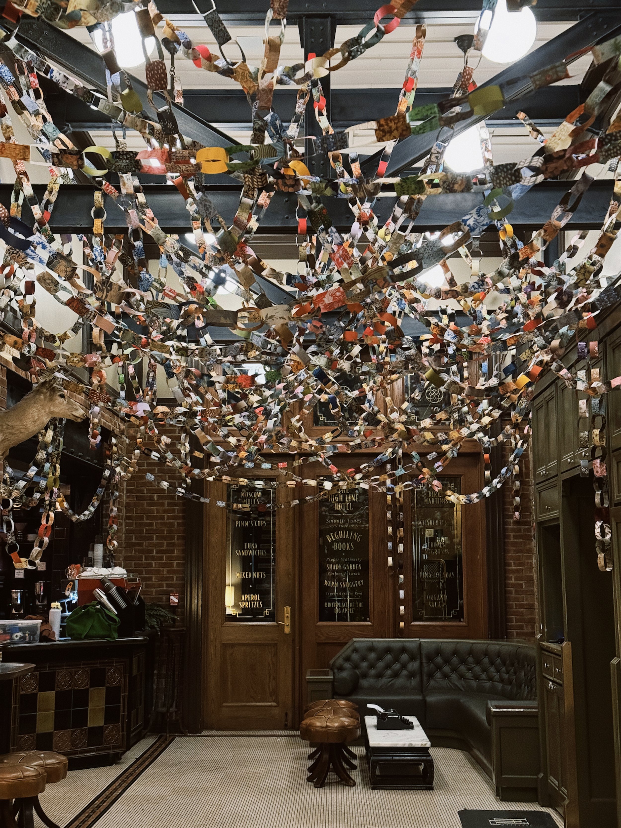 Indoor cozy space decorated with overlapping paper chains hanging from the ceiling and a vintage style interior with brick walls, wooden furniture, a leather tufted corner bench, and a small round leather stool. There is a door with chalkboard signage and a counter with coffee-making equipment.