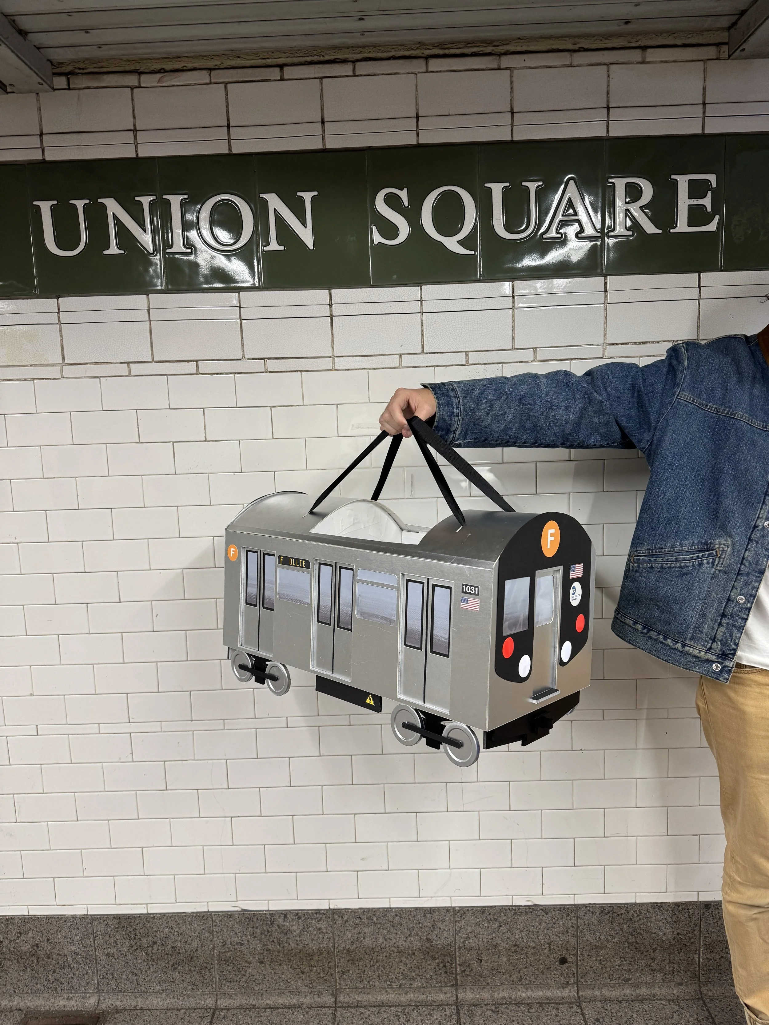 Person holding a subway train Halloween costume in front of a subway station sign.