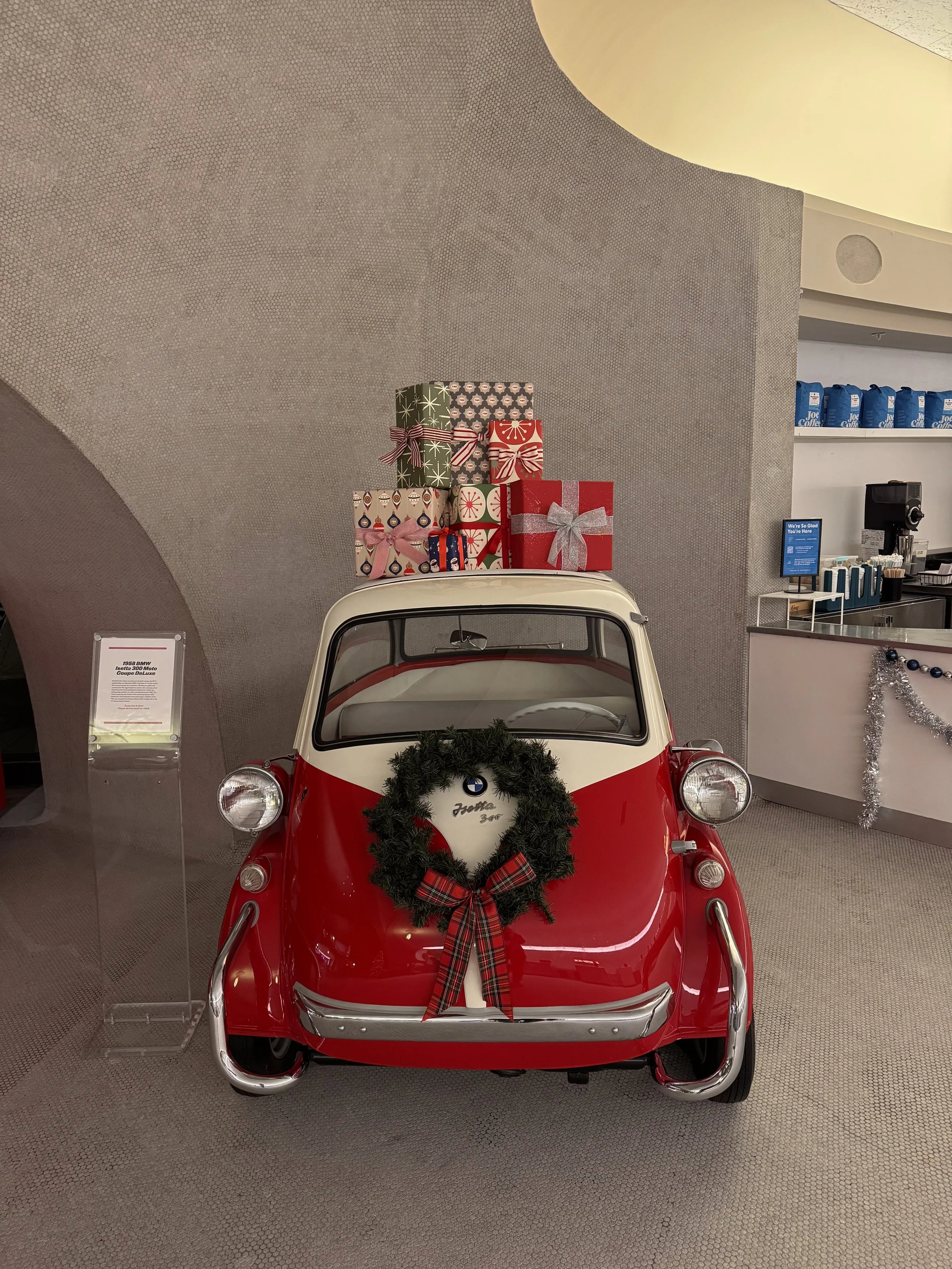 A small red and white vintage BMW Isetta with a Christmas wreath on its front and a stack of wrapped Christmas presents on its roof, displayed in a holiday-themed setting.