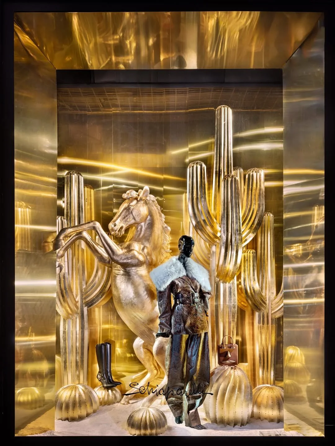 Elegant retail display with gold background featuring a rearing gold horse sculpture, tall gold cactus structures, black cowboy boots, a luxurious coat with white fur collar, a handbag, and gold decorative elements on the floor.