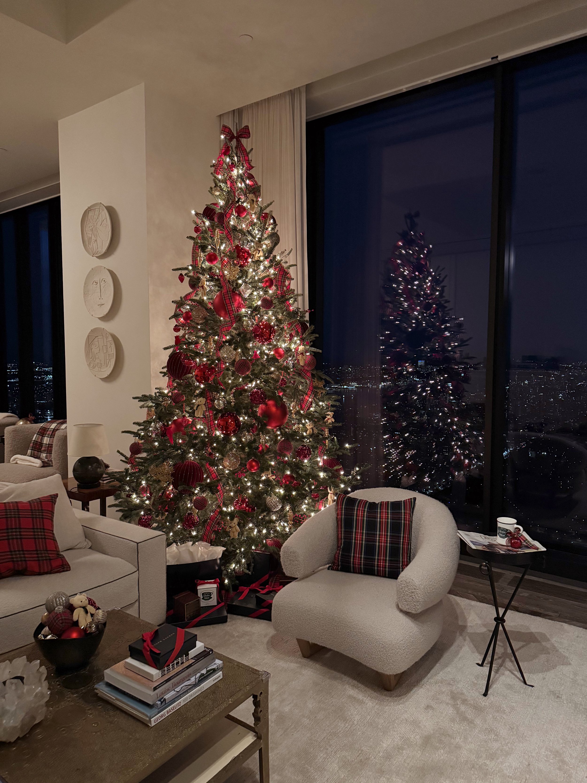 A decorated Christmas tree with red ornaments and ribbons in a living room, with a large window reflecting the tree at night.