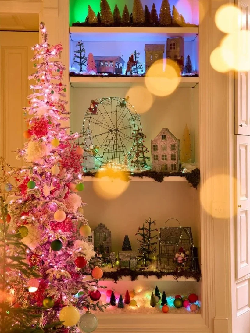 A Christmas display on shelves with a pink decorated Christmas tree on the left, miniature trees, buildings, a Ferris wheel, and figurines decorated with holiday lights and ornaments.