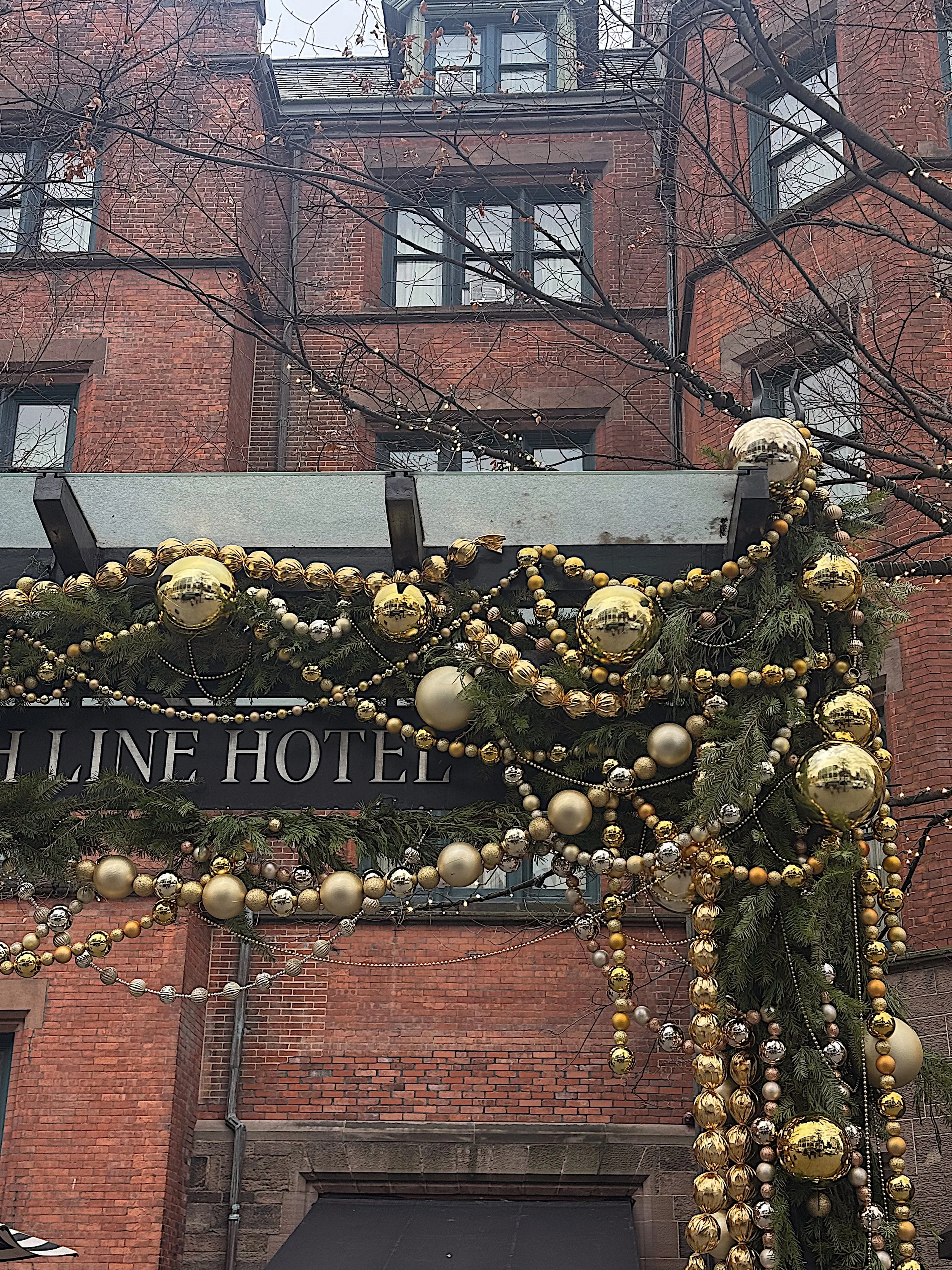 Holiday decorations with gold and silver ornaments, greenery, and string lights on a hotel sign, in front of a red brick building with trees in the background.