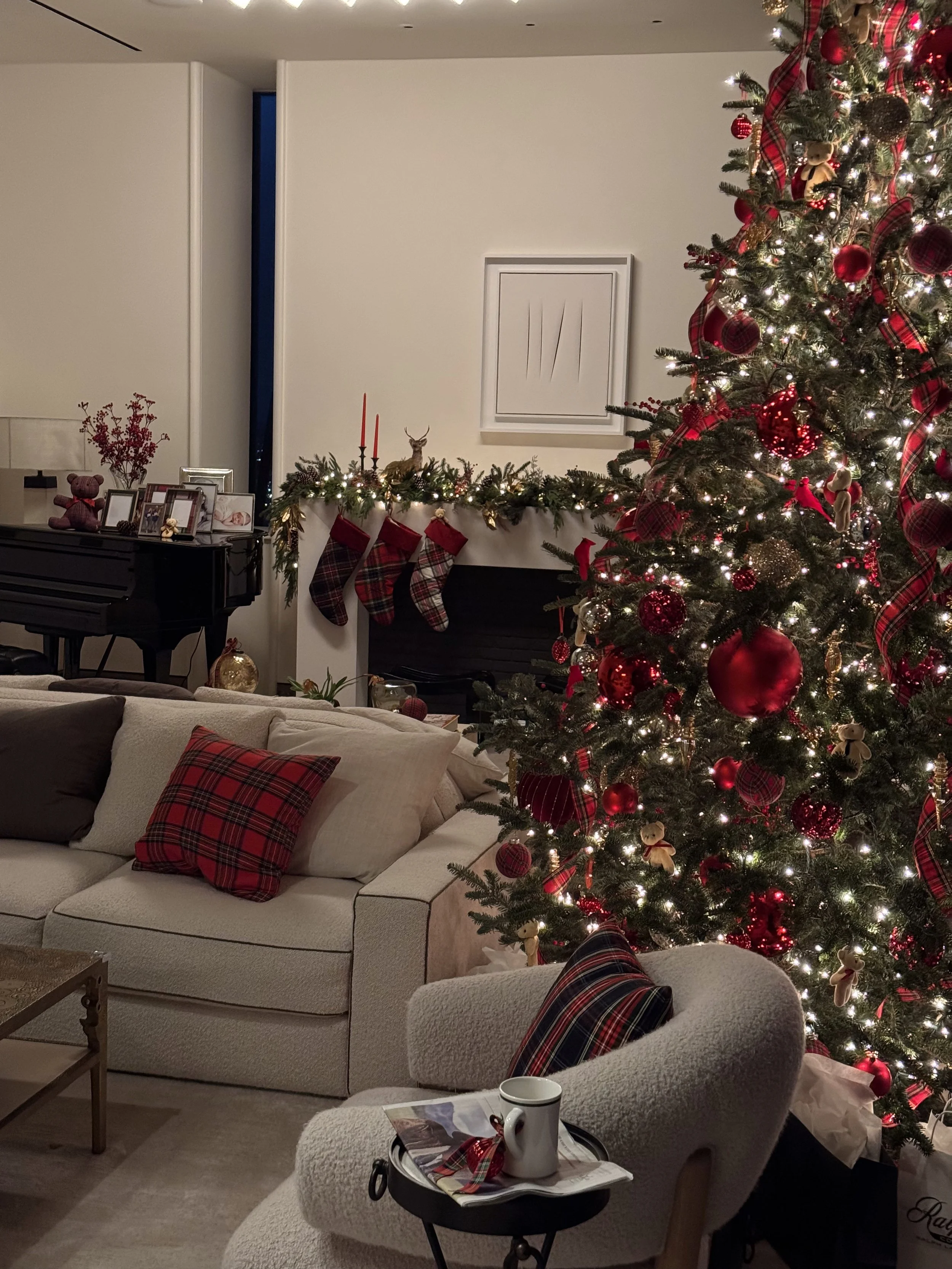 Christmas decorated living room with a large decorated Christmas tree, a sofa with plaid and neutral pillows, a cozy armchair with a plaid pillow, a fireplace mantle with stockings, garland and candles, a piano with photos and decorations, and a side table with a mug, magazines, and a key.