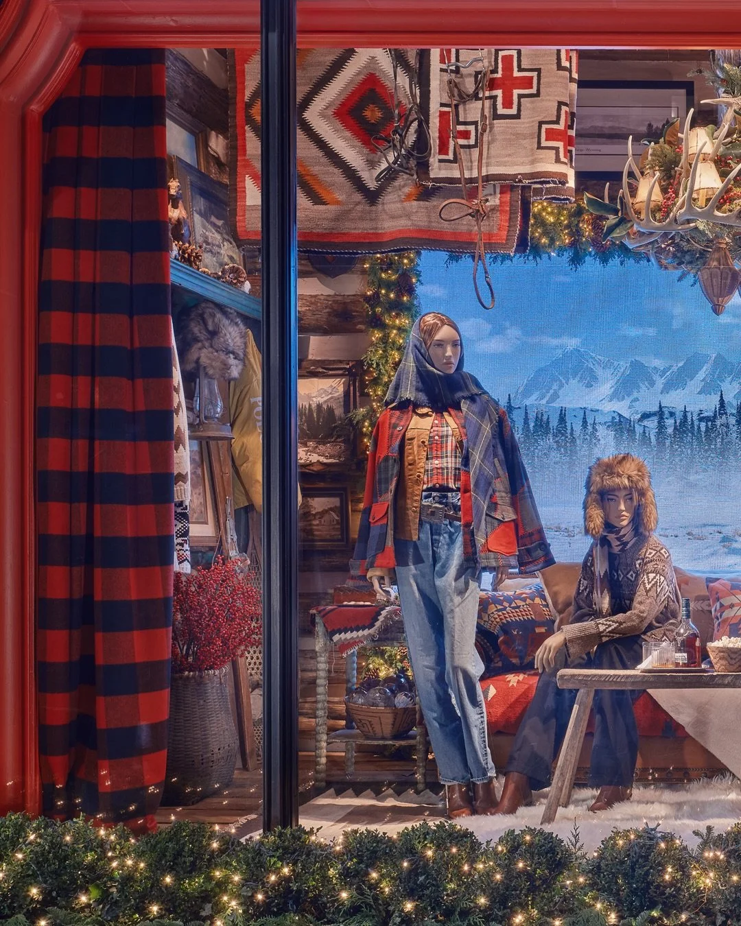 Winter-themed storefront display with mannequins dressed in plaid and winter clothing, surrounded by holiday decorations, rugs, and a snowy mountain backdrop.