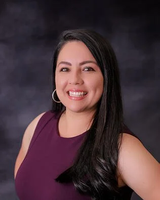 A smiling woman with long dark hair wearing a sleeveless purple top and hoop earrings, standing against a dark background.