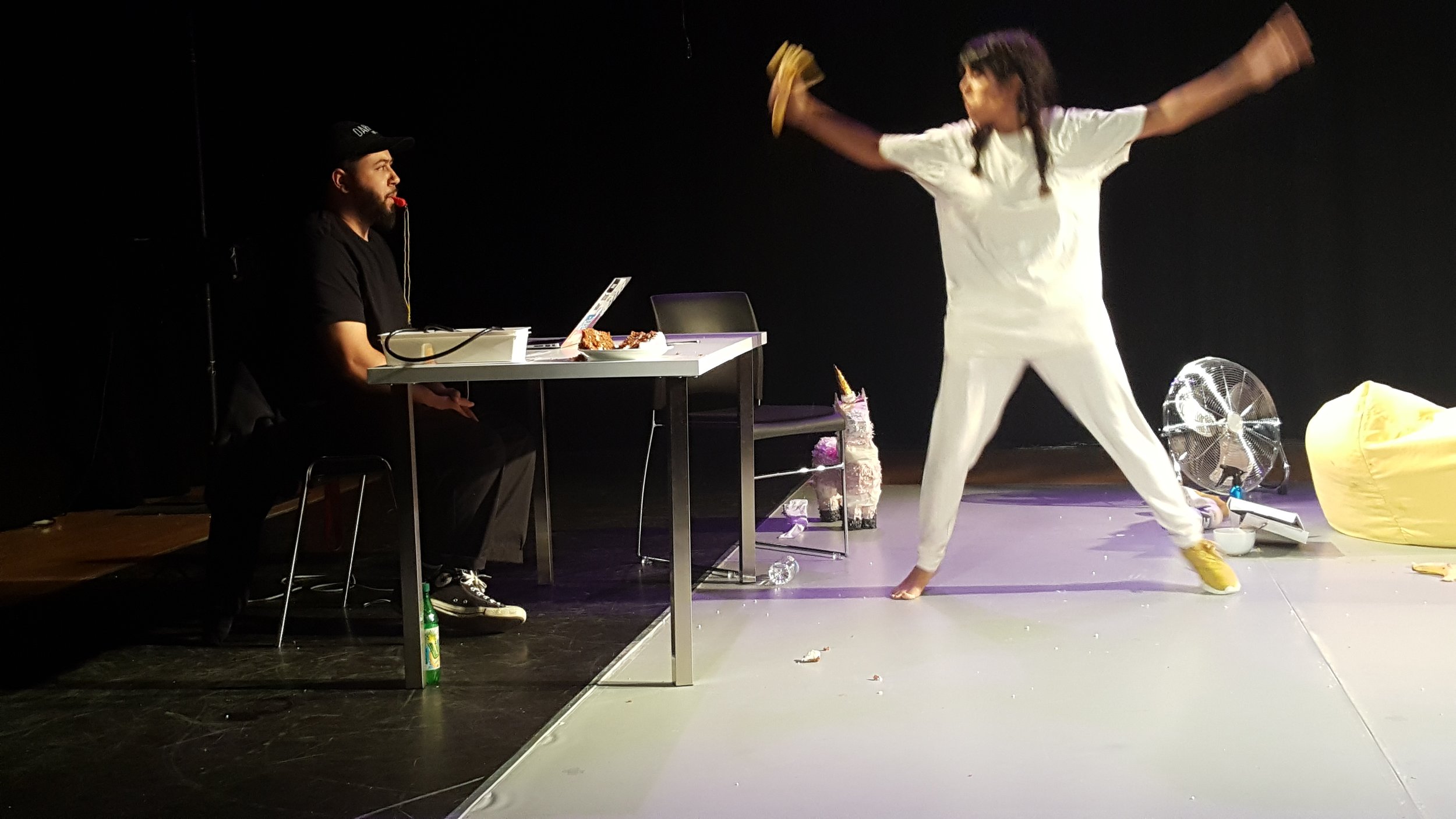  Image description: Man sits at a table on stage with a whsitle in mouth looking at a woman who is throwing a banana towards him.  