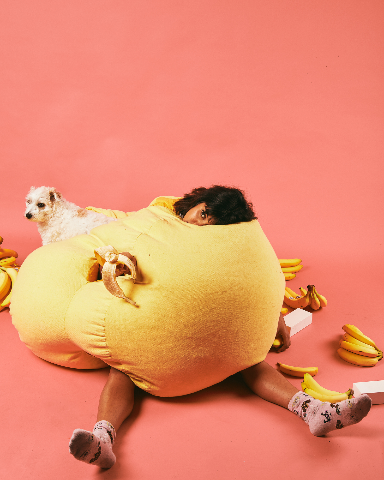  Woman sits in a yellow beanbag in a pink backdrop. A white dog is at the back. 