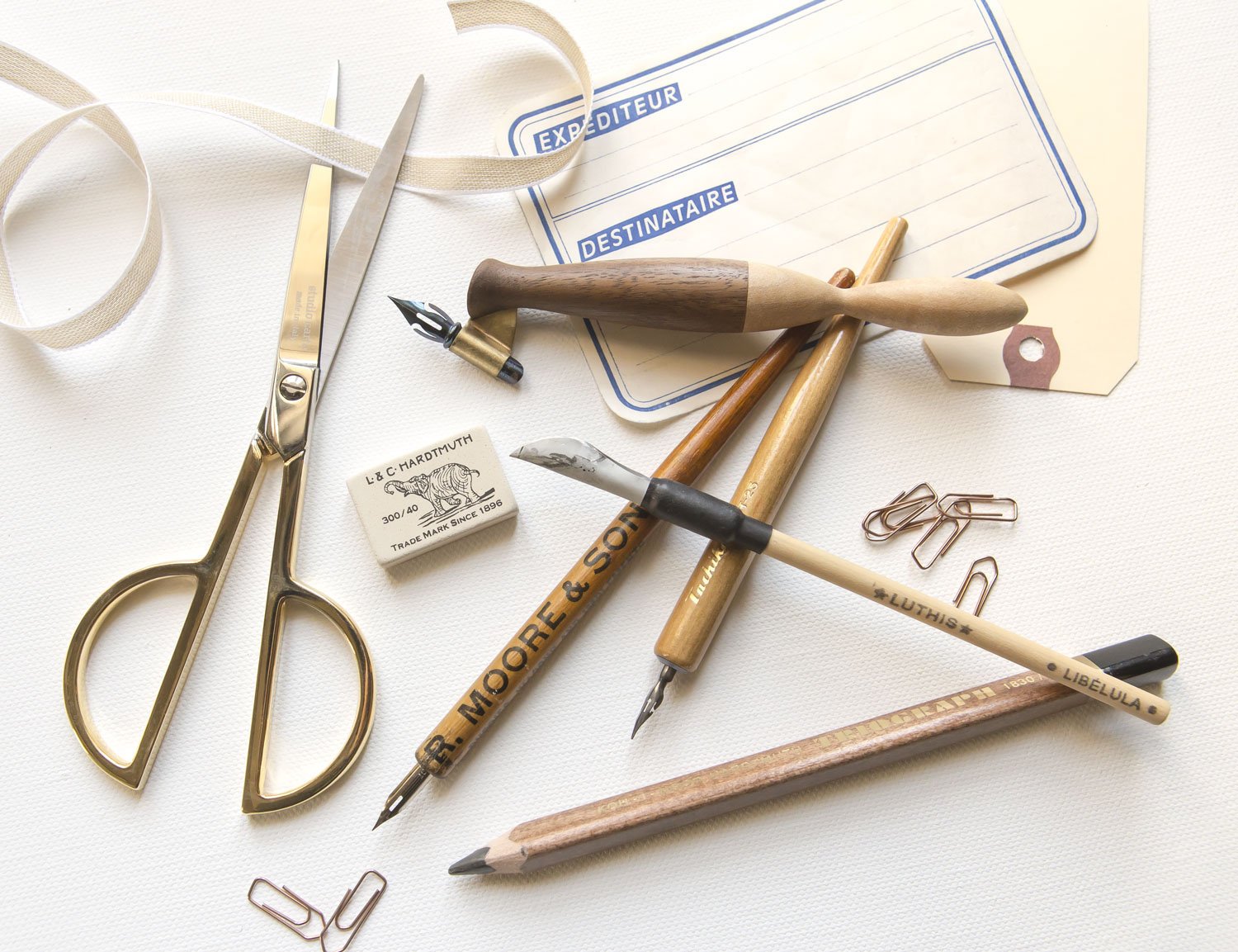 A collection of vintage calligraphy and desk tools, including scissors, pointed pens pens, pencils, paperclips, labels, and a ribbon, arranged on a white surface.