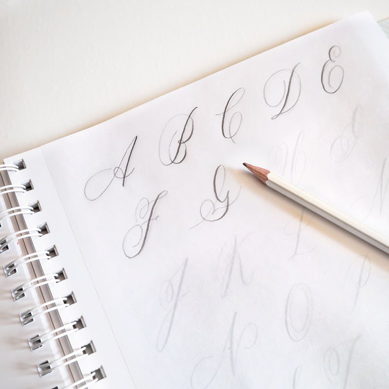 Tracing paper with handwritten flourished capital letters and pencil lying on top of an open book page from Mastering Modern Calligraphy by Molly Suber Thorpe