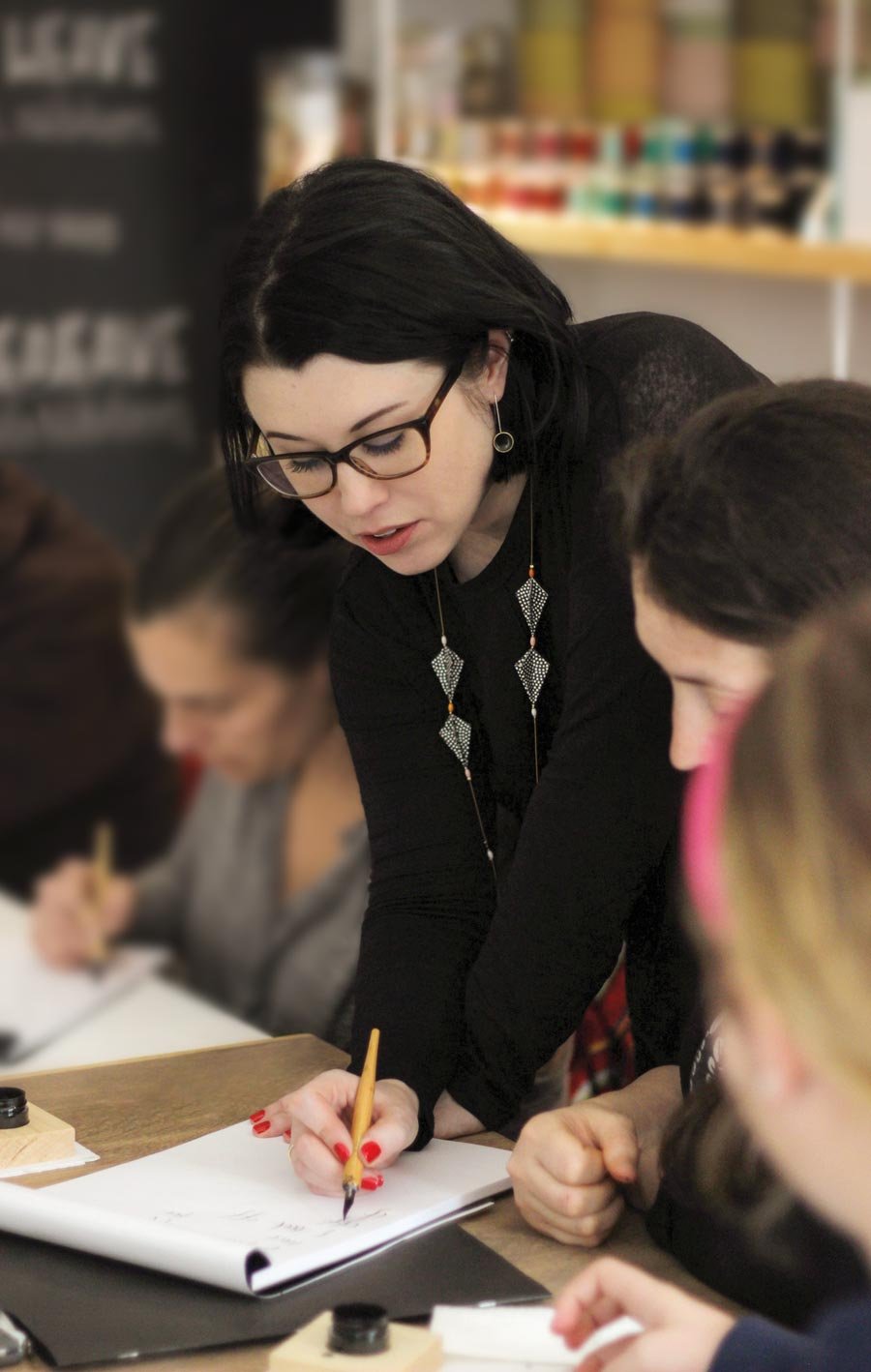 Molly Suber Thorpe teaching a modern calligraphy workshop, learning over a student’s paper helping them form letters with a pointed calligraphy pen and black ink.