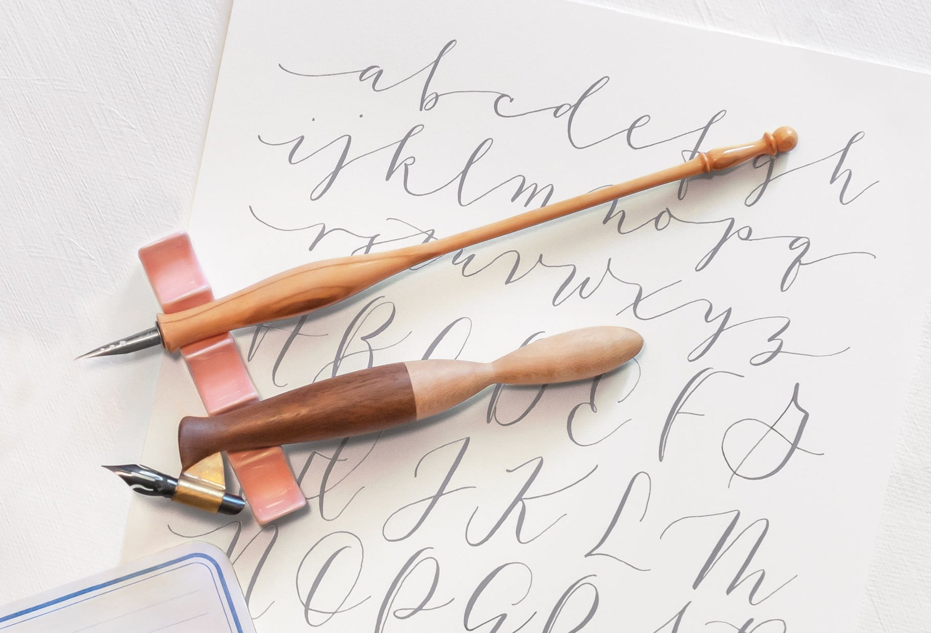 A modern calligraphy alphabet written in gray gouache ink on white paper, with two wooden calligraphy pens and a pink ceramic pen rest