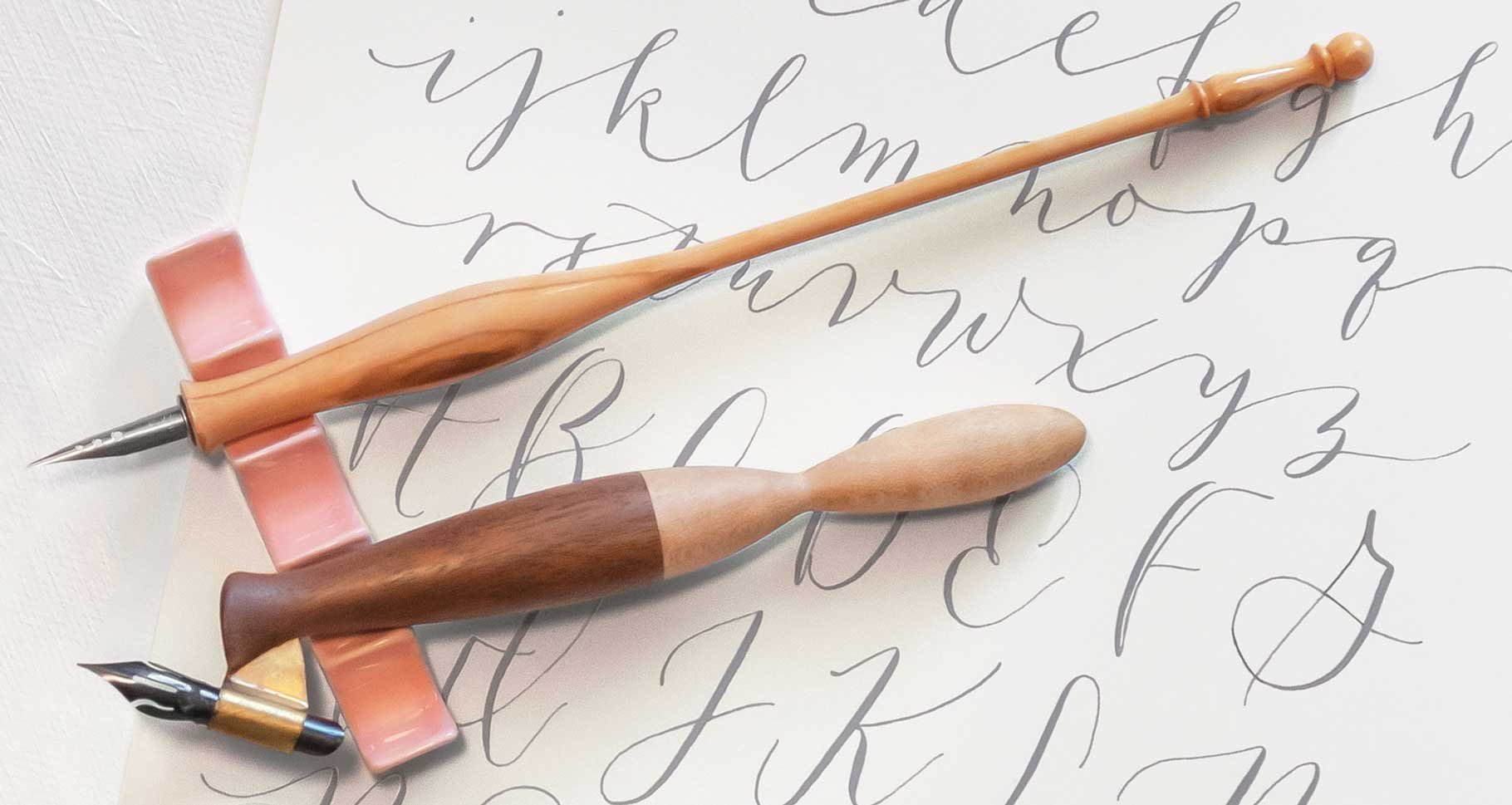 Modern calligraphy alphabet in gray ink on white paper, with two wooden calligraphy pens resting on a pink ceramic pen rest