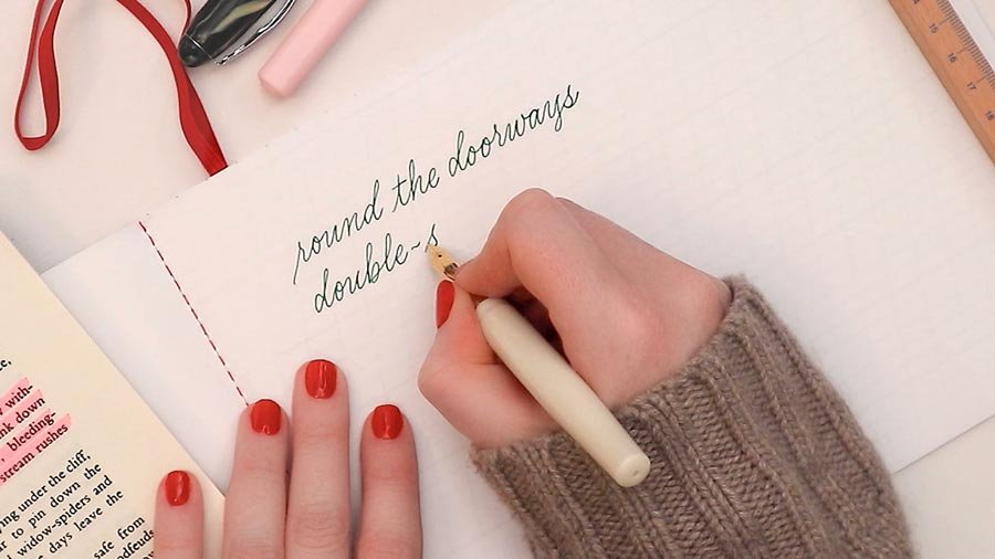 Molly Suber Thorpe with red painted fingernails writing in a notebook with a white fountain pen and green ink; the unfinished text says “round the doorways, double…“ There are open books, a ruler, and writing tools on the surrounding desk.
