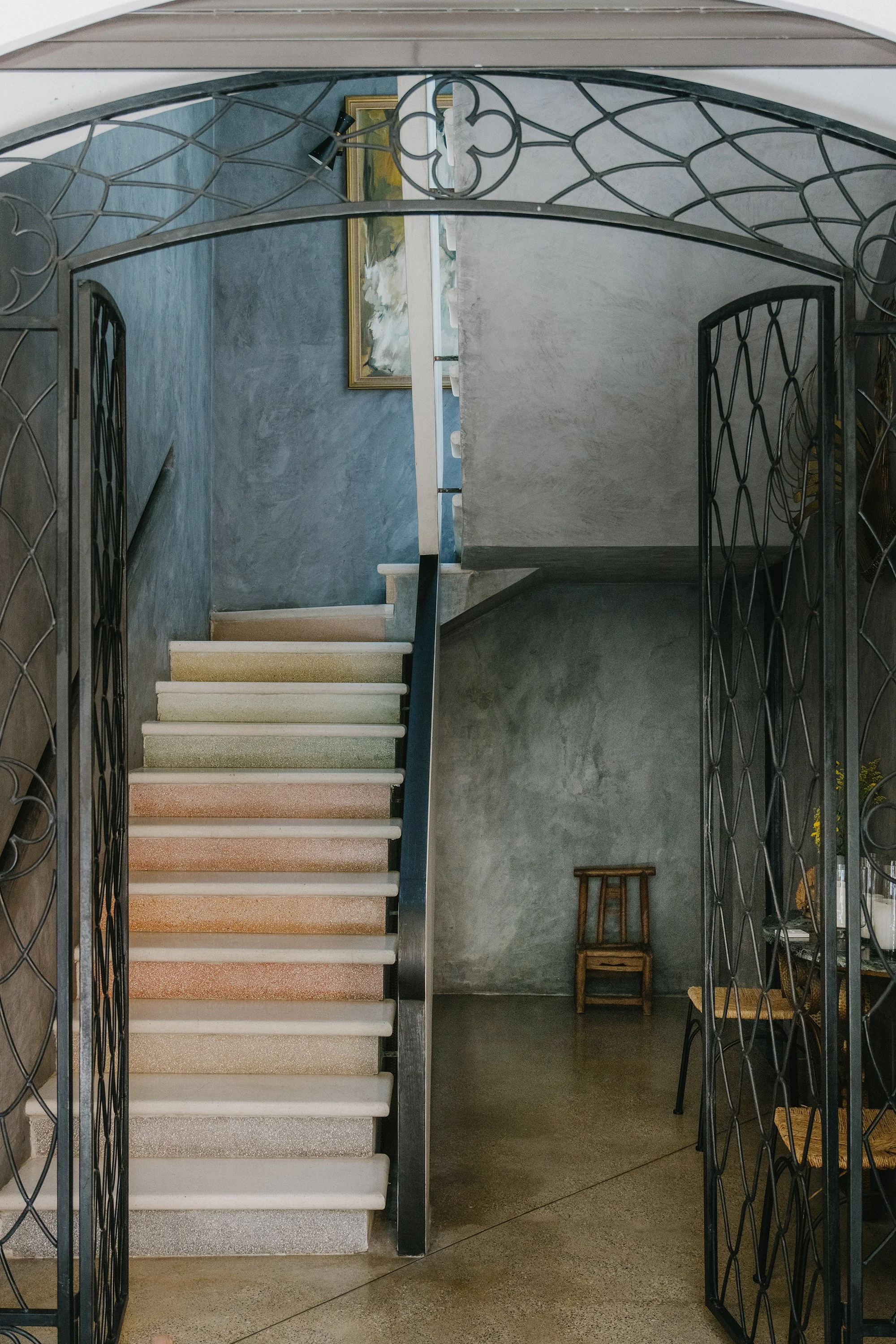 Interior view of a staircase with a metal railing on either side. The stairs have light-colored risers and darker treads. In the background, there is a blue textured wall, a painting in a gold frame, and a small stool. Mundos Interiores. Ciudad Colon