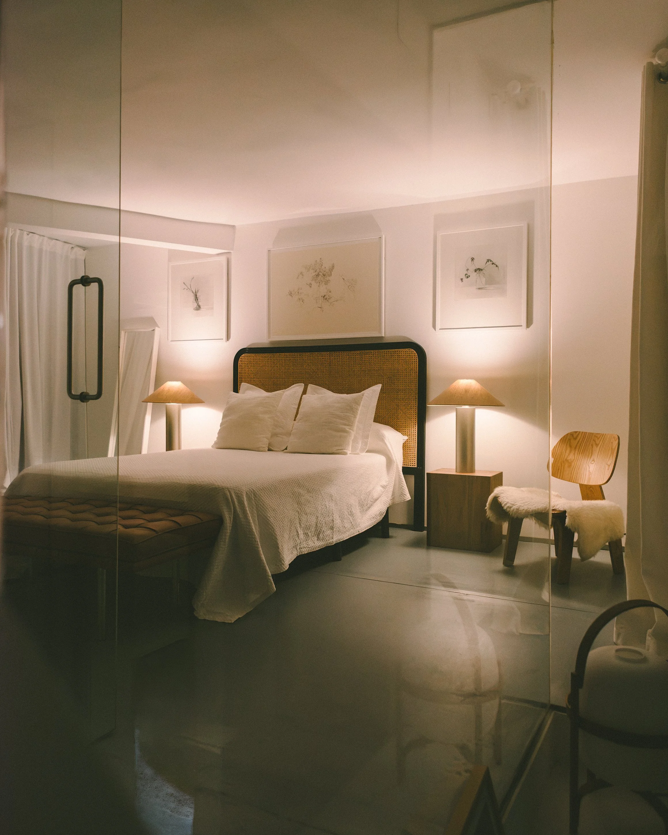 A bedroom seen through a glass door, with a bed, pillows, bedside lamps, and framed artwork on the wall.
