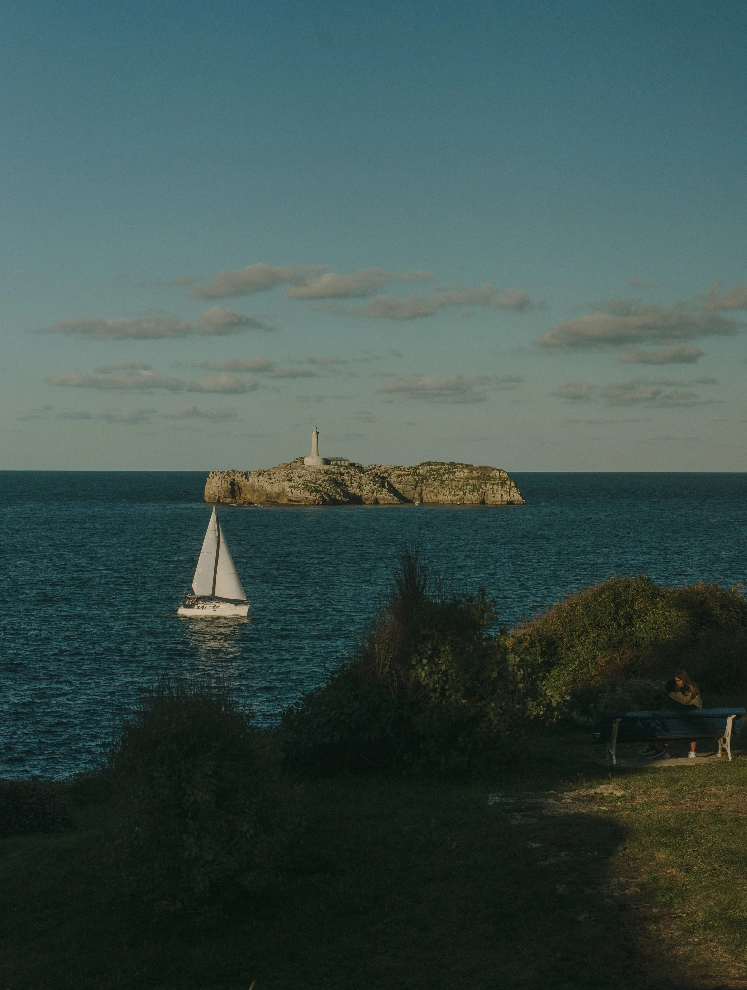 An ocean scene with a sailboat in the foreground, an lighthouse on a rocky island in the middle ground, and a partly cloudy sky in the background.