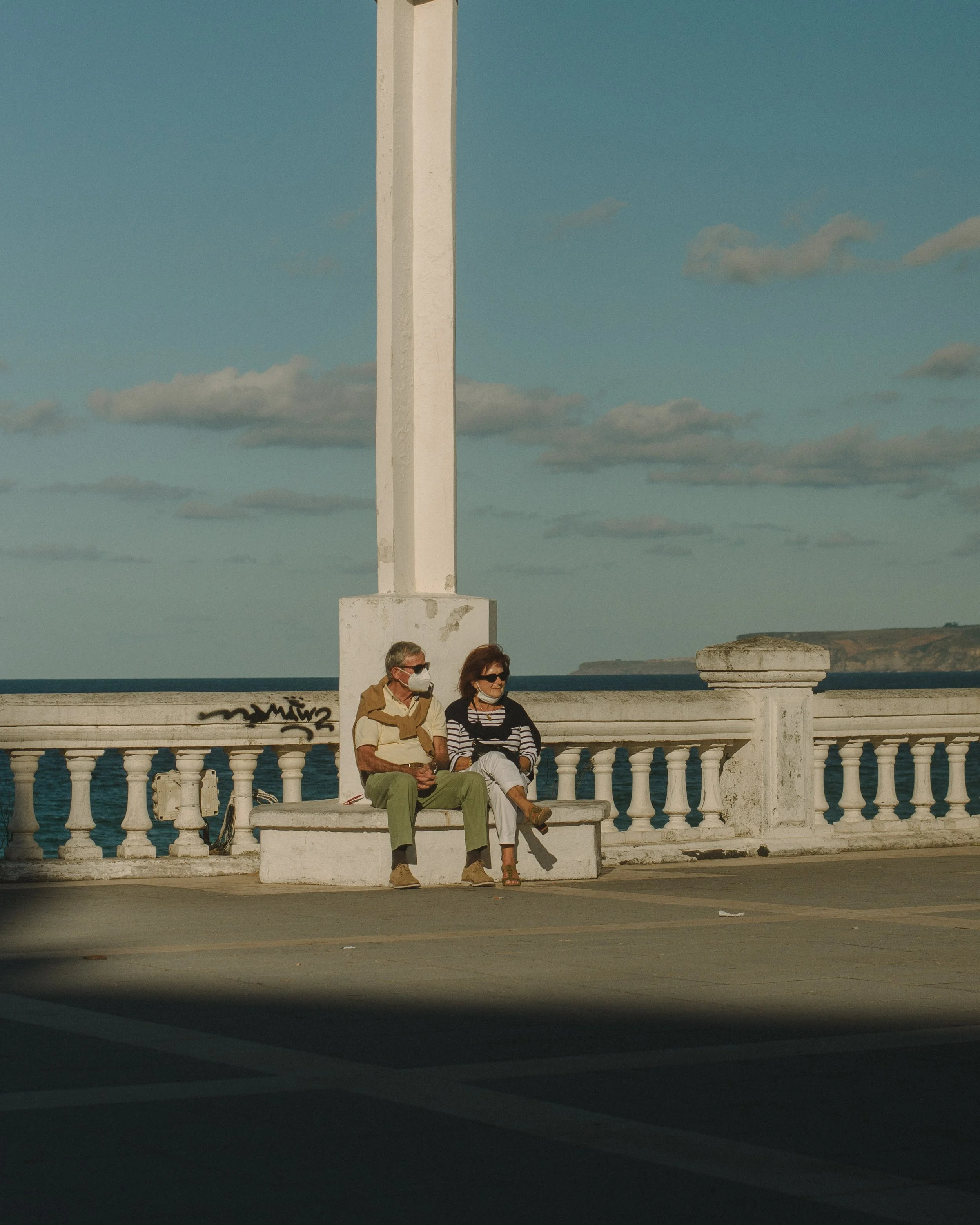 Two people sitting on a white bench near the seaside, wearing face masks, with a tall white monument behind them and a clear blue sky with scattered clouds above.