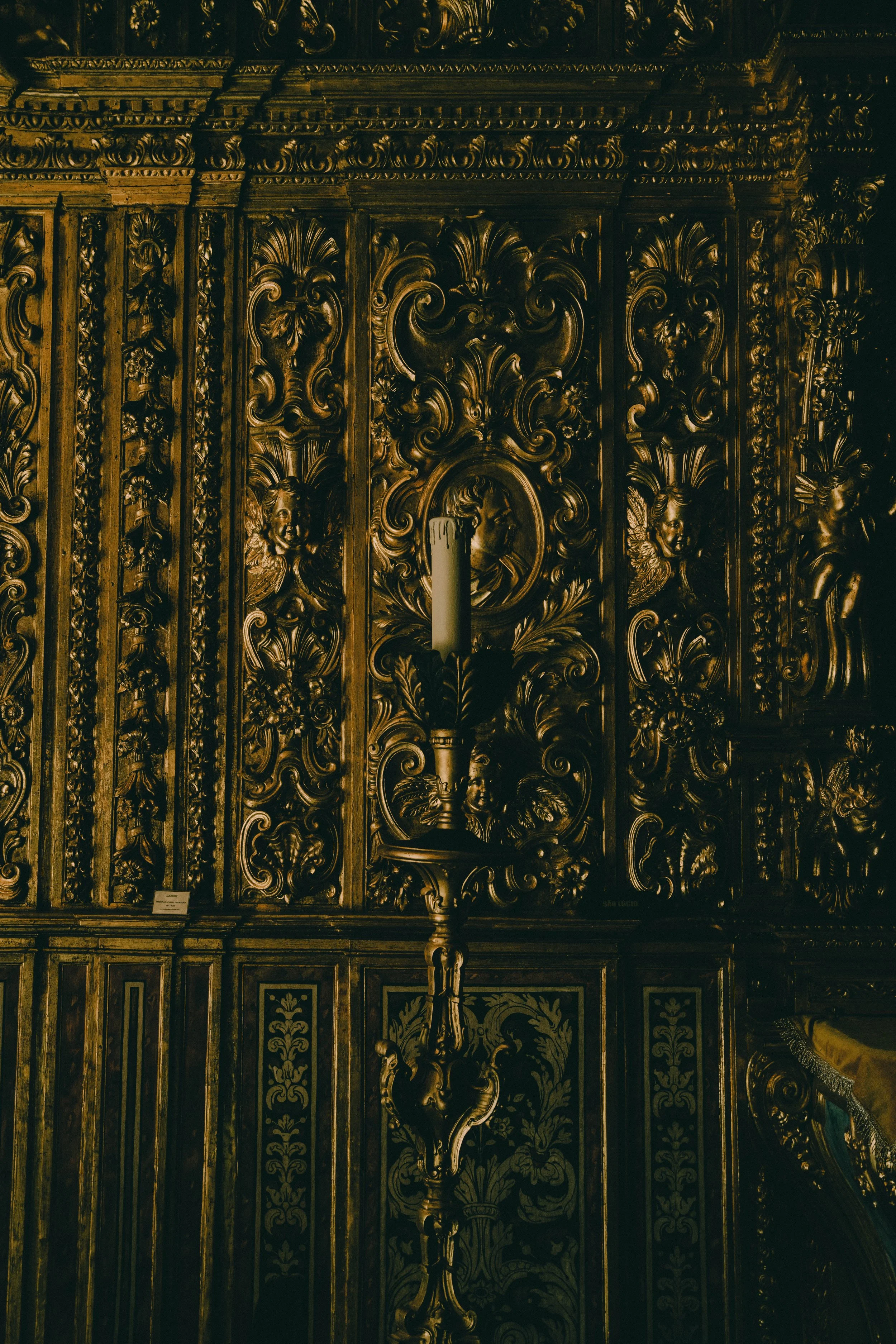 An ornate, gilded, carved wooden panel with intricate floral and cherub motifs, featuring a candle holder with a candle.
