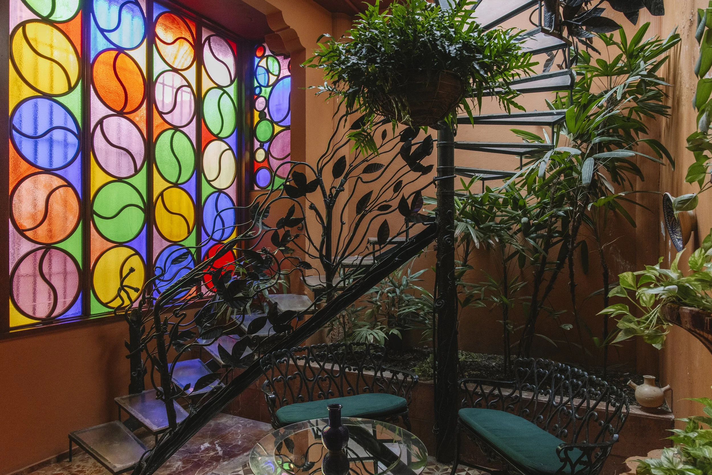 Indoor area with a brightly colored stained glass window featuring large, overlapping circles in red, orange, yellow, green, blue, and pink. There are various green plants and a decorative metal spiral staircase in the foreground. A small glass table