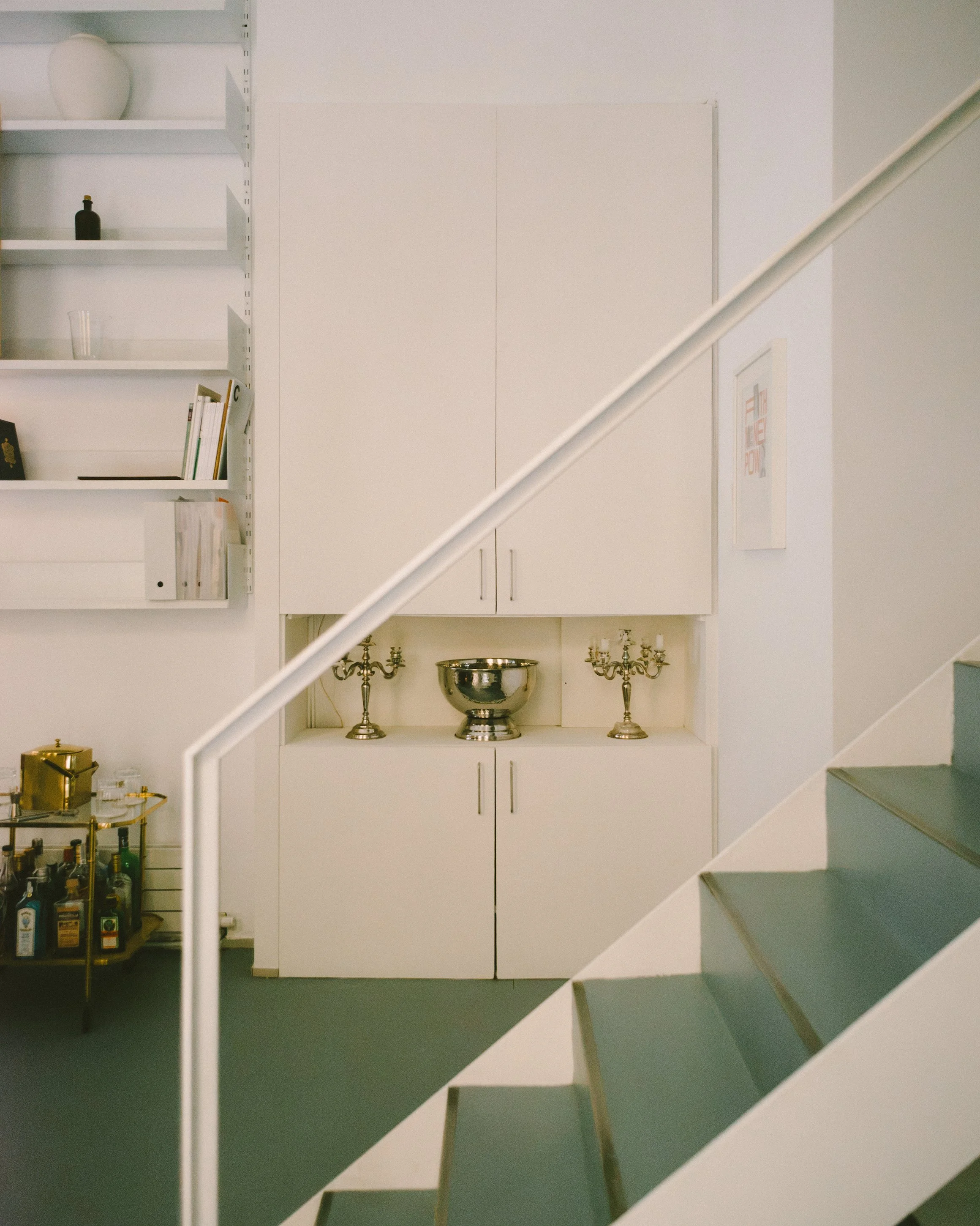 Interior view of a room with white cabinets and shelves, a silver bowl, candelabras, and framed artwork on the wall. The photo is taken from the bottom of a staircase, partially obscuring the view.
