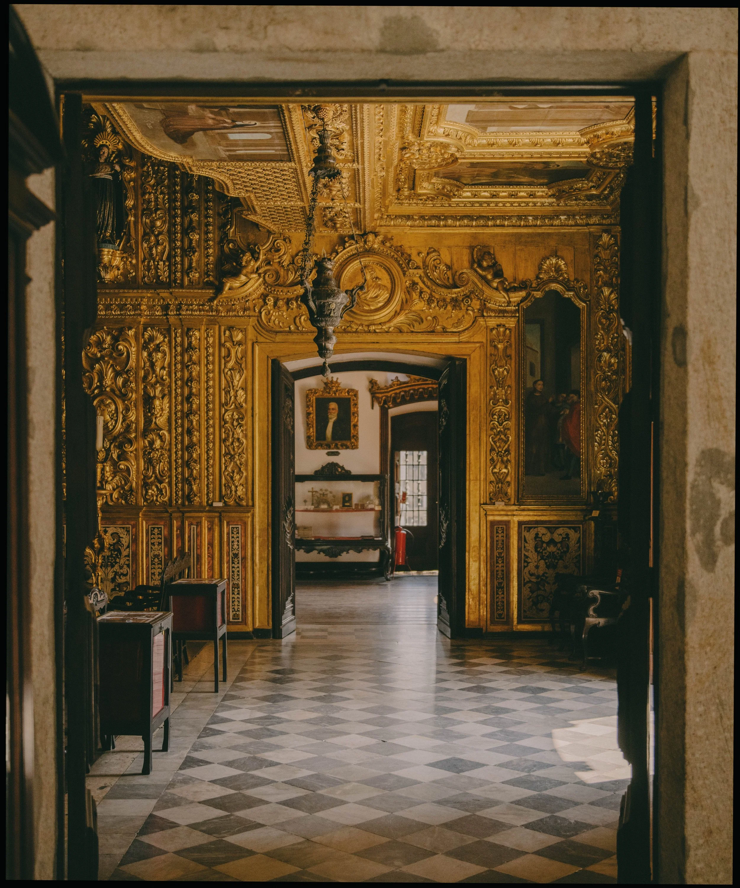 An ornate hallway with golden wood carvings, paintings, and chandeliers in a historic building, viewed through a doorway.