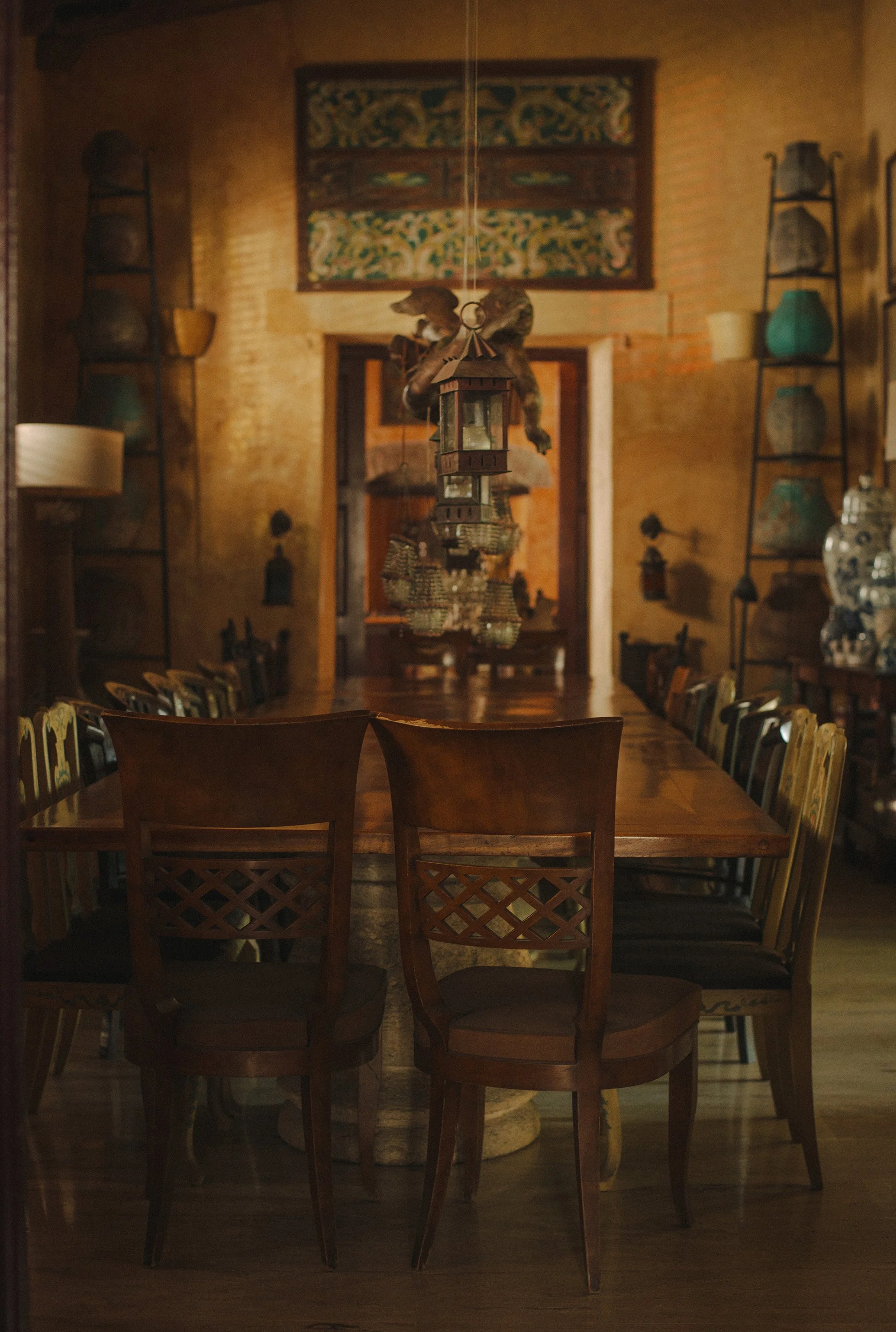 A dimly lit dining room with a long wooden table and eight wooden chairs. A decorative lantern chandelier hangs above. Shelves with pottery and artwork on the walls. Mundos Interiores. Ciudad Colonial de Santo Domingo.