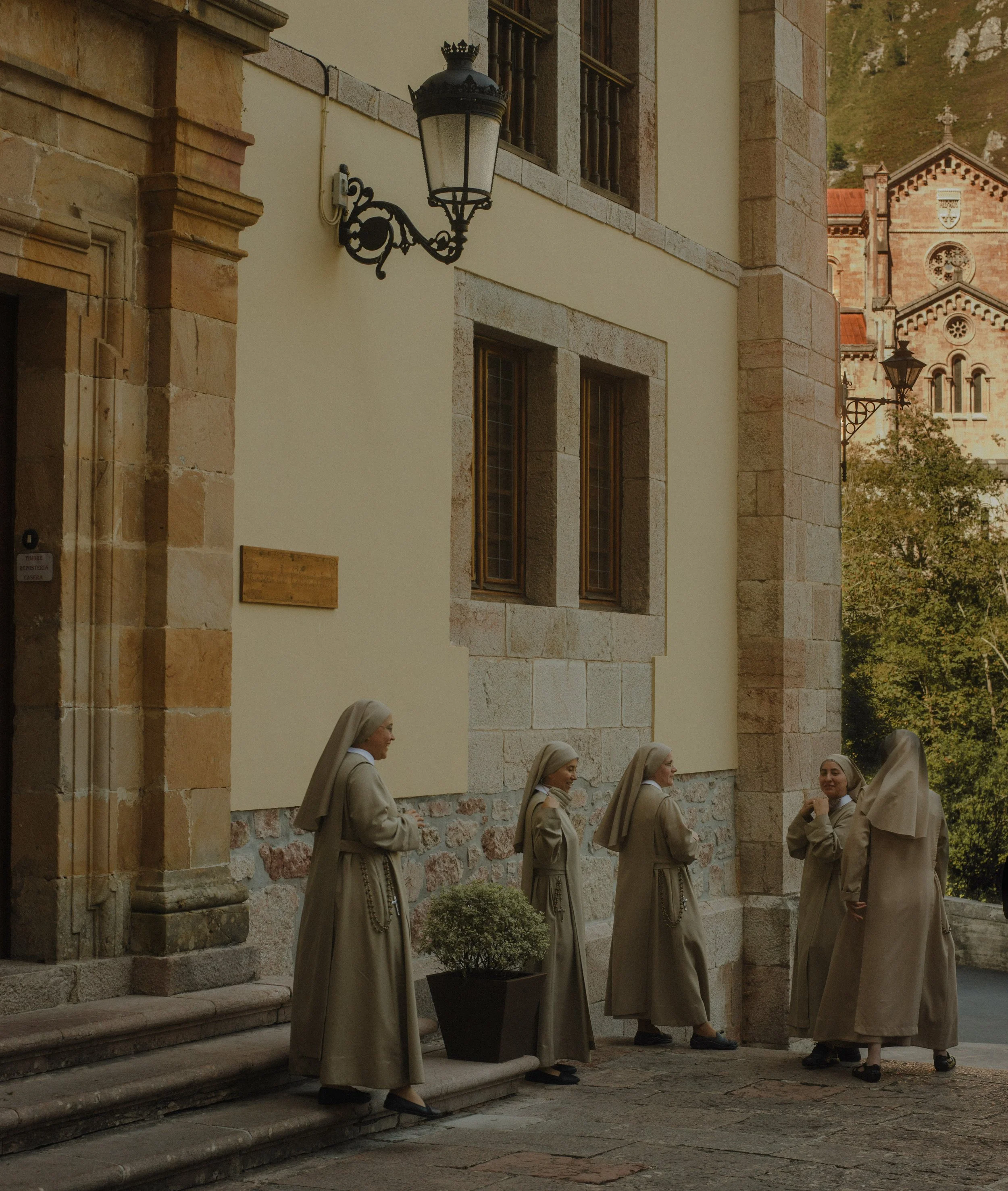 A group of nuns dressed in beige habits standing in a line outside a stone and plaster church, engaged in conversation. The church has stone archways, windows, and decorative lamp posts.